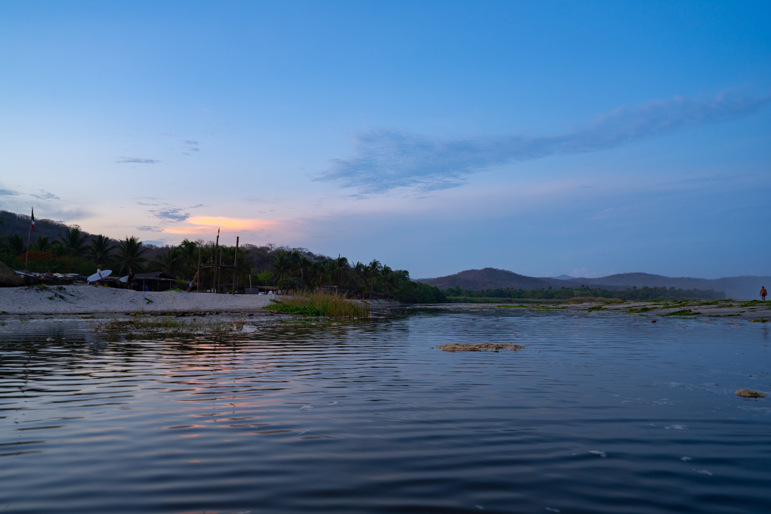 A river flowing through a landscape with a sky filled with clouds at sunset, and some trees and structures along the banks.