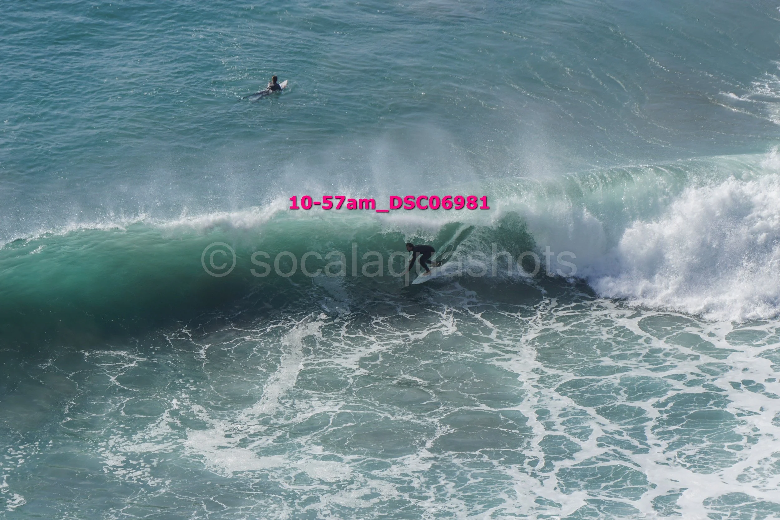 A person surfing on a large wave in the ocean, with another surfer in the background.