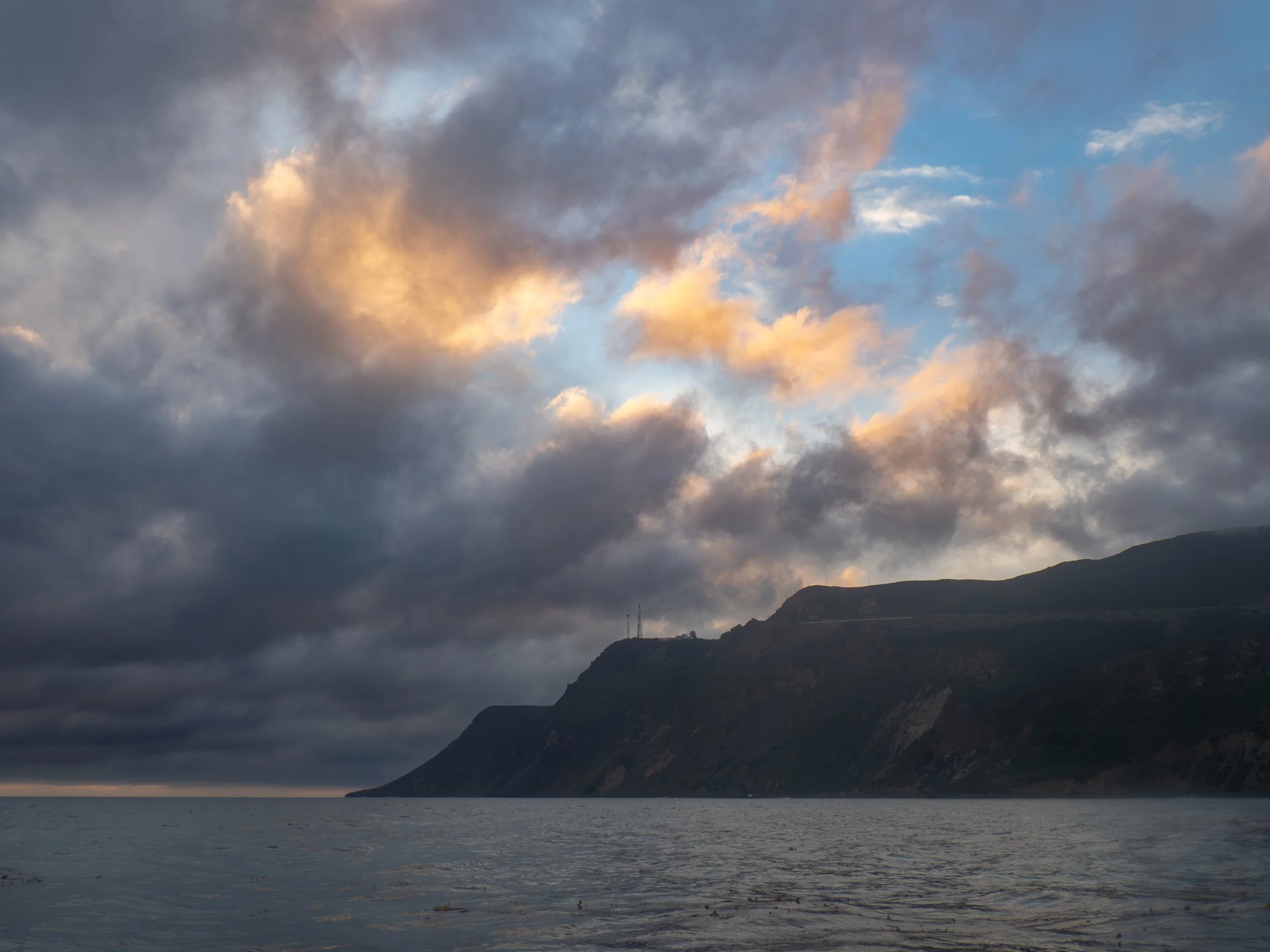 A view of a large body of water near dark hills or cliffs under a cloudy sky with patches of sunlight