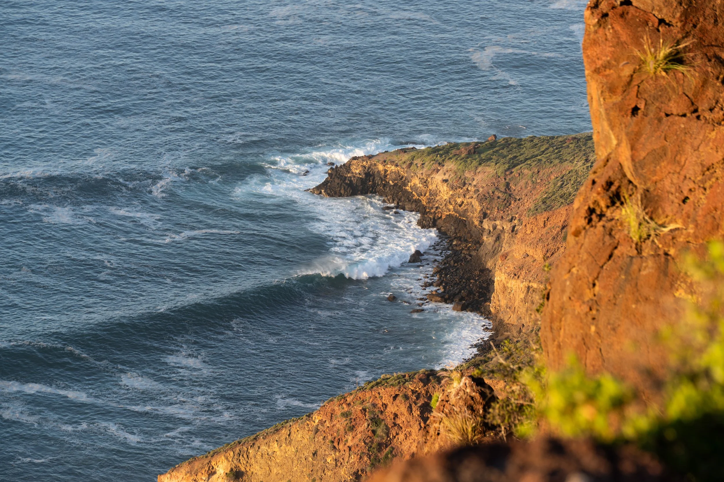 A rugged coastline with orange-brown cliffs and ocean waves crashing against the rocks at sunset.