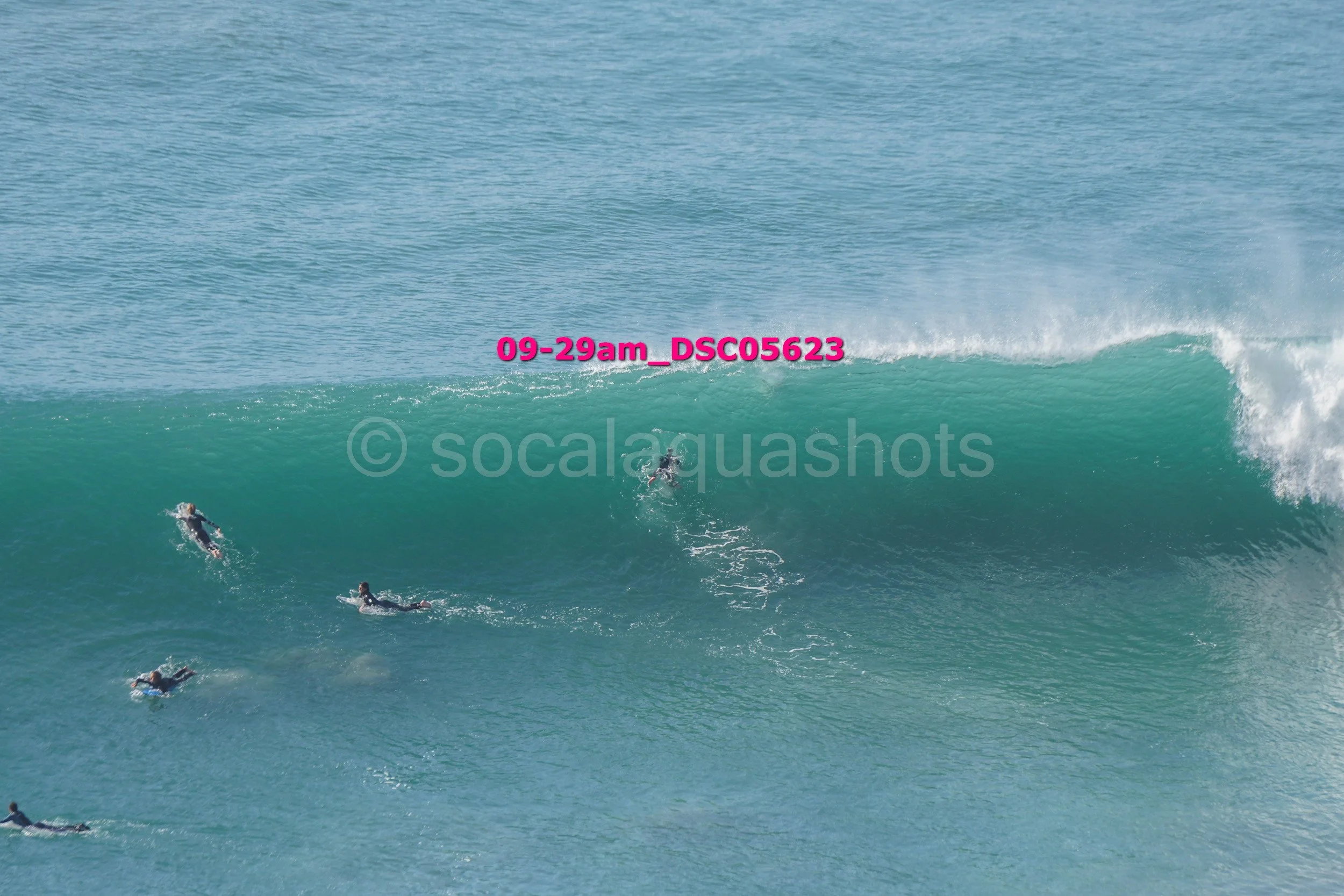 Surfers riding a large ocean wave with clear blue water and a partly cloudy sky in the background.