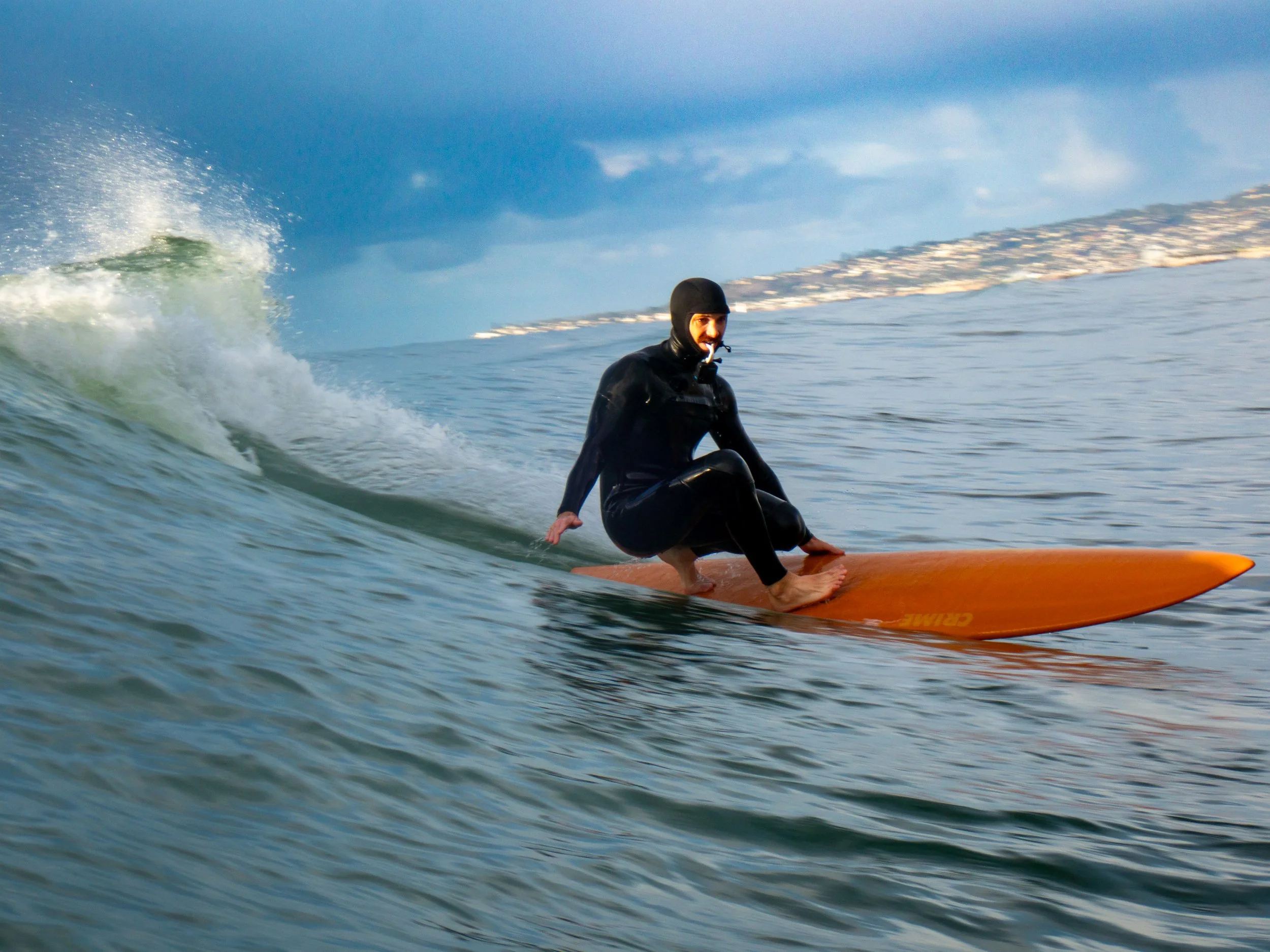 A person wearing a wetsuit and helmet surfing on an orange surfboard in the ocean with a distant landmass and cloudy sky in the background.