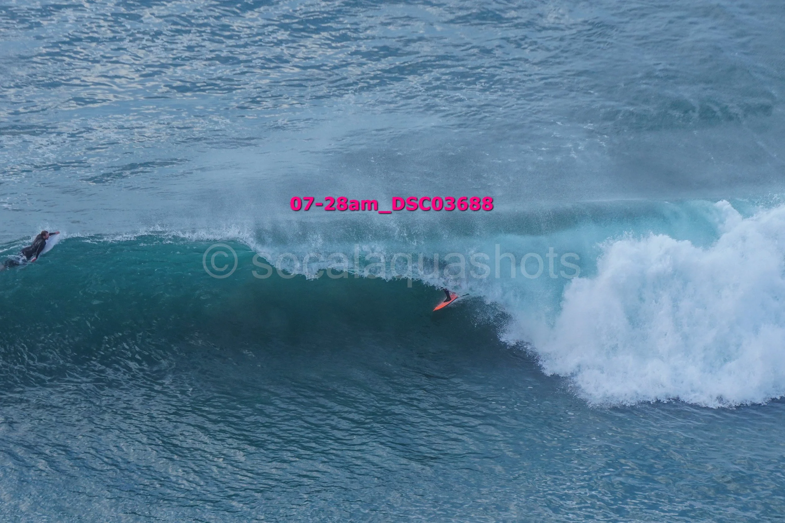 A surfer riding a wave in the ocean, with another surfer visible in the water nearby.