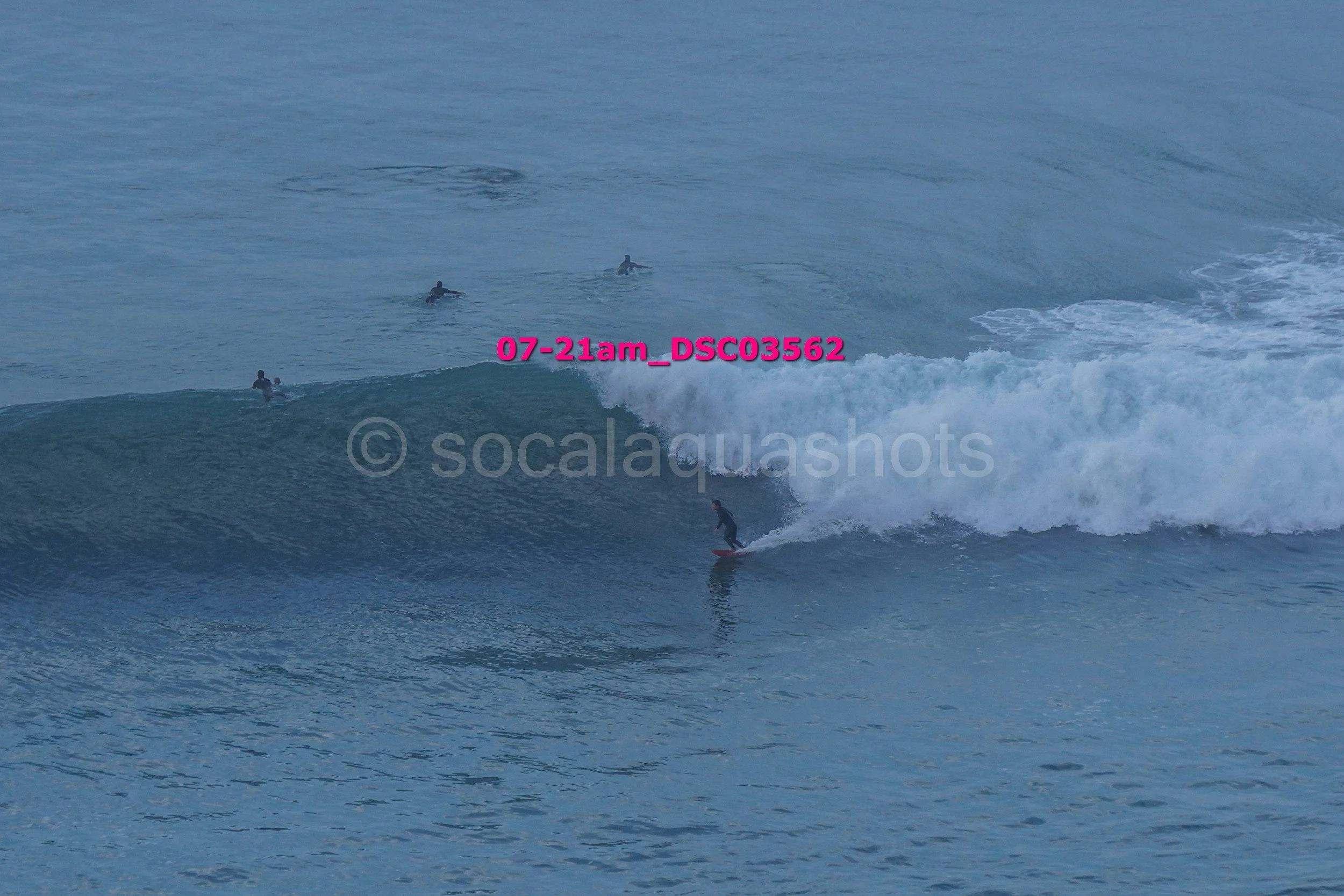 A person surfing on a wave in the ocean, with several swimmers or surfers in the background.