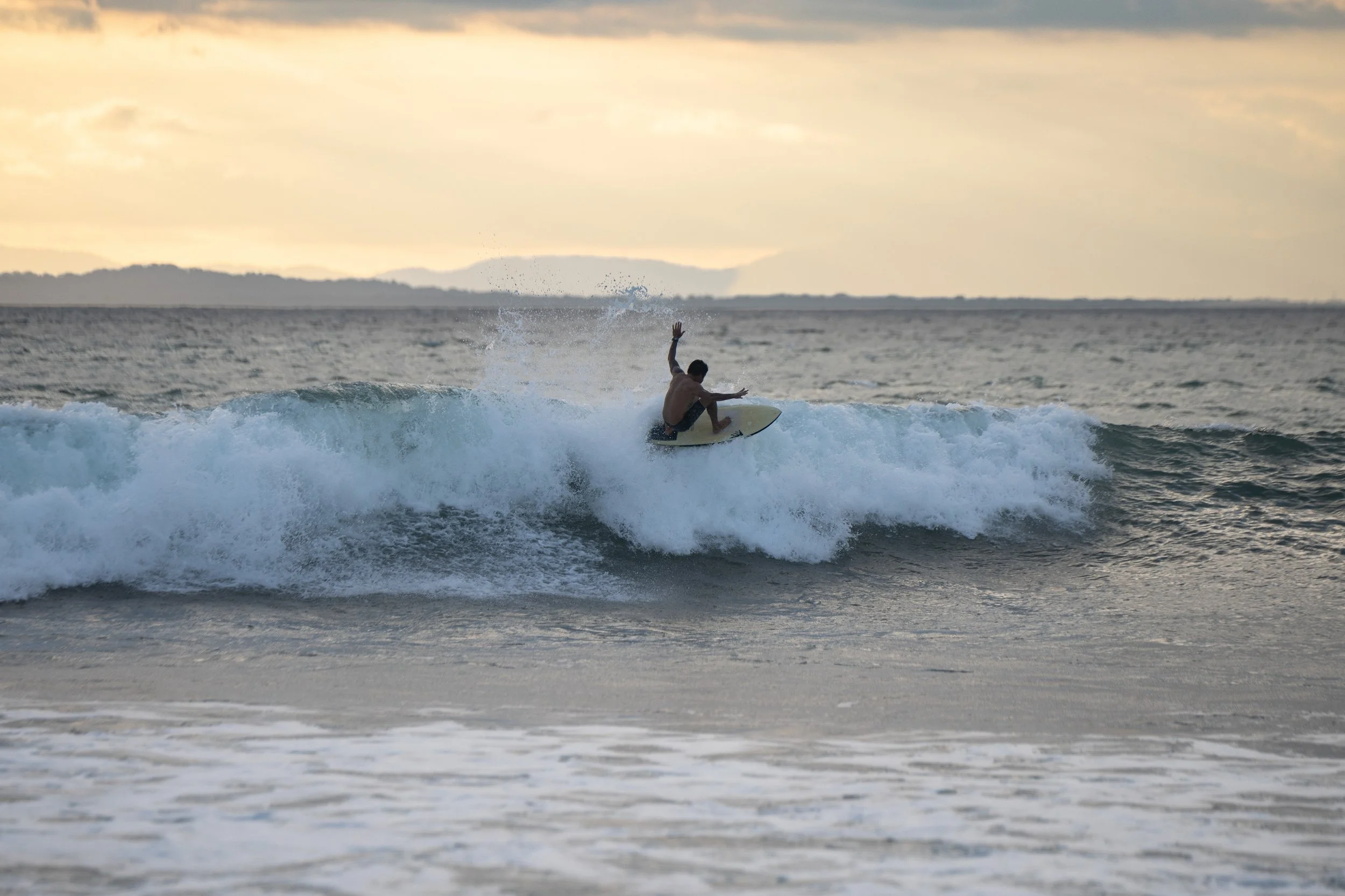A person surfing on a wave in the ocean during sunset or sunrise, with a distant shoreline and cloudy sky.