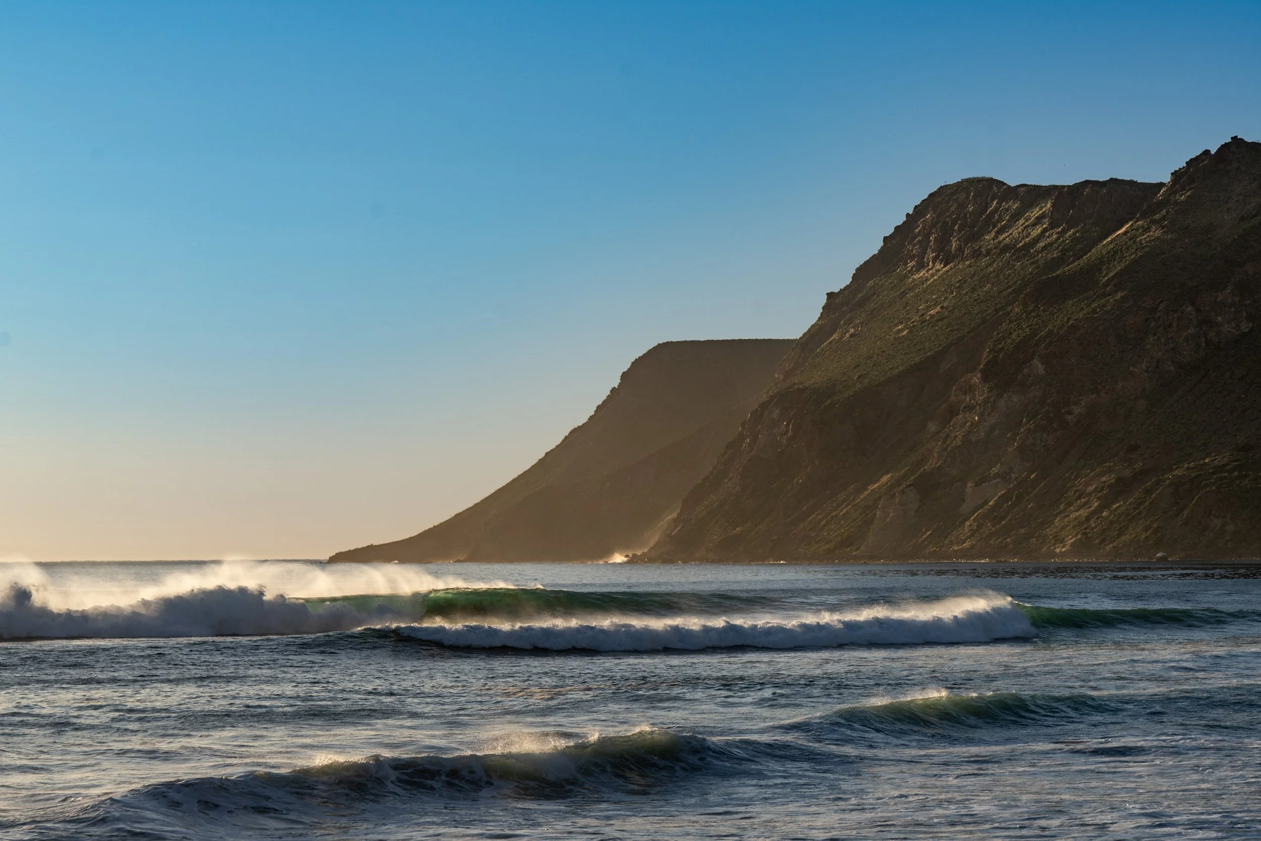 Sunset over the ocean with waves crashing and rugged cliffs in the background.