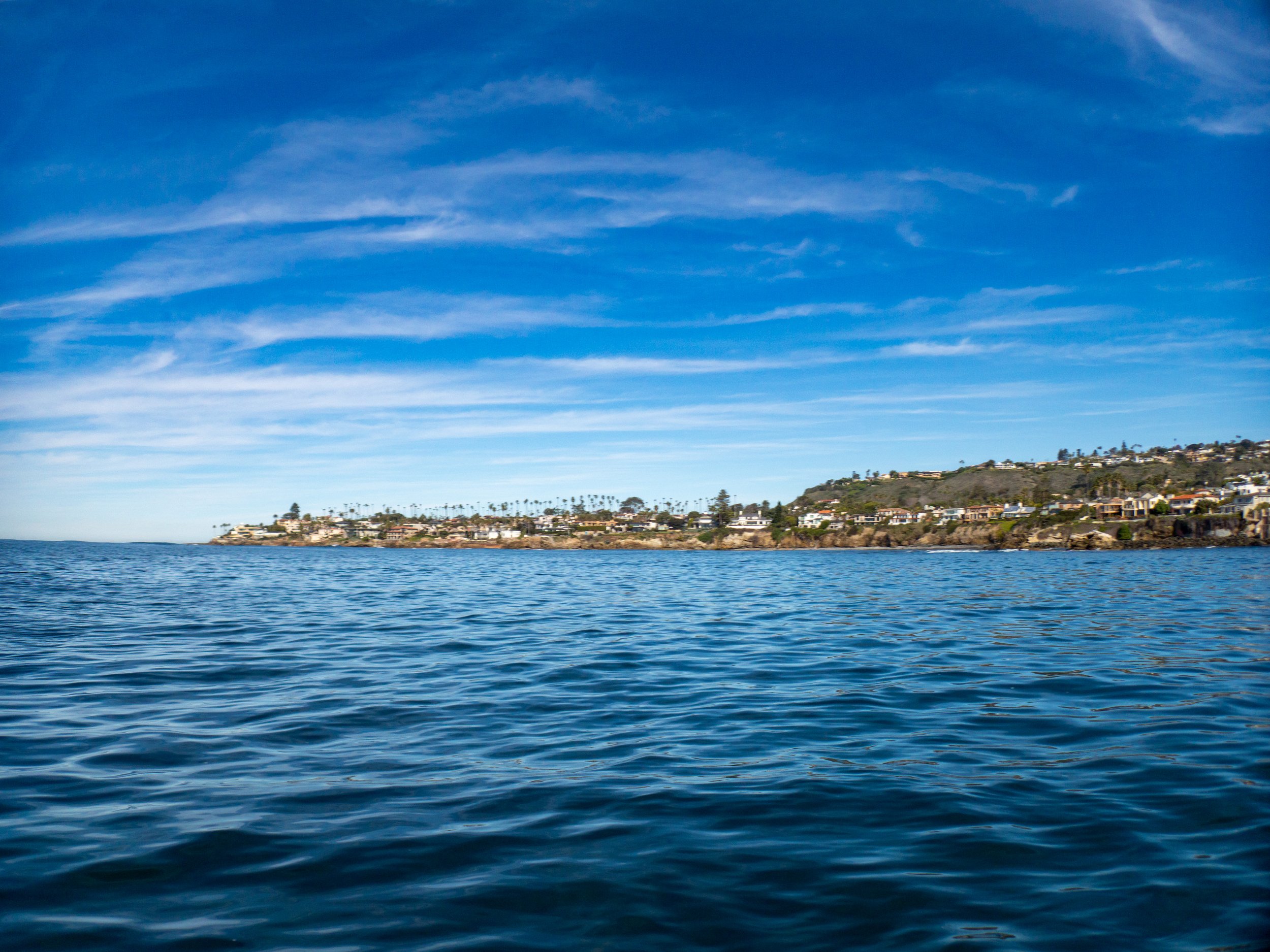 View of a coastal area with houses and palm trees on a hillside, seen across calm ocean waters with a vibrant blue sky and streaks of clouds.