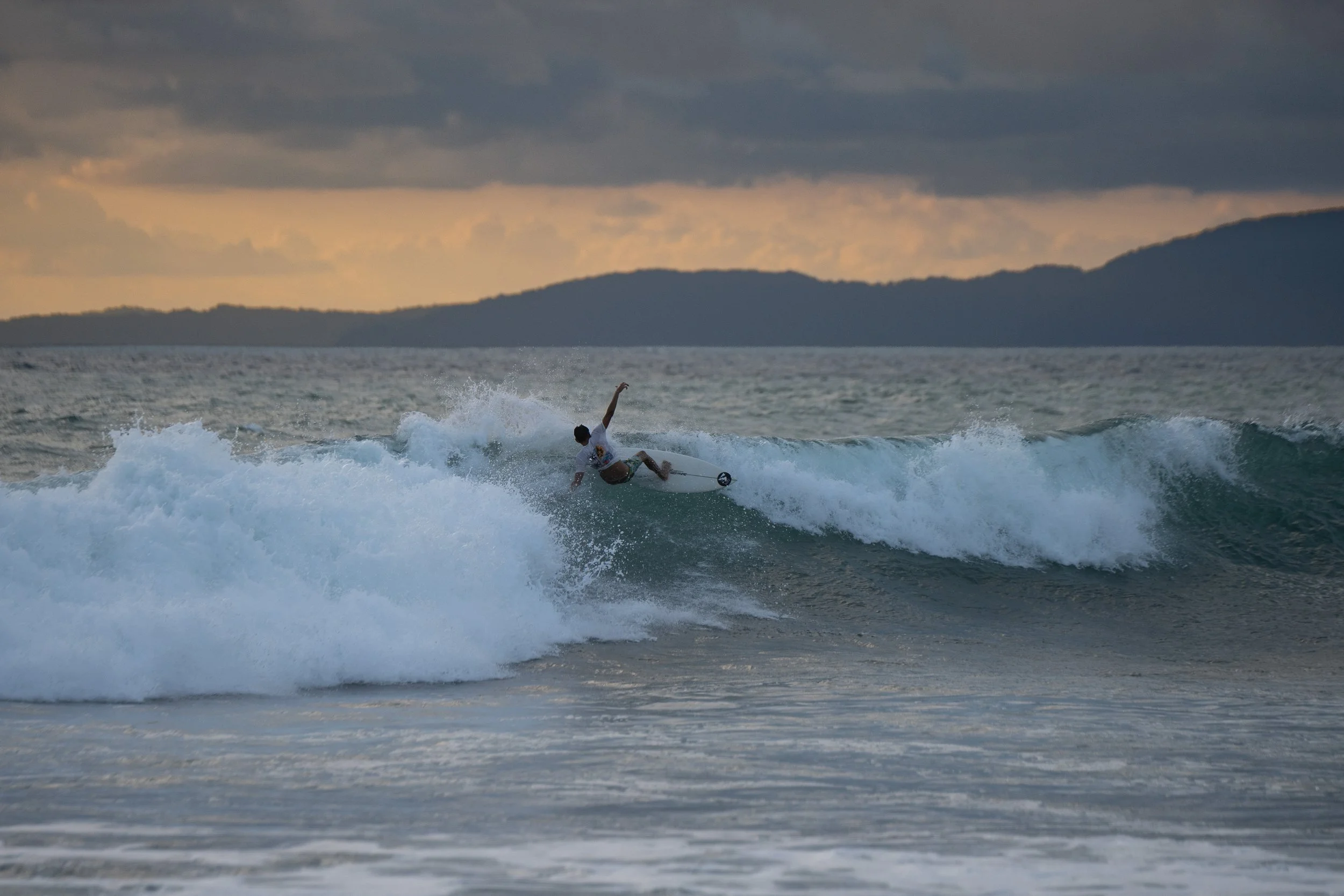 A person riding a wave on a surfboard in the ocean during sunset, with mountains and cloudy sky in the background.