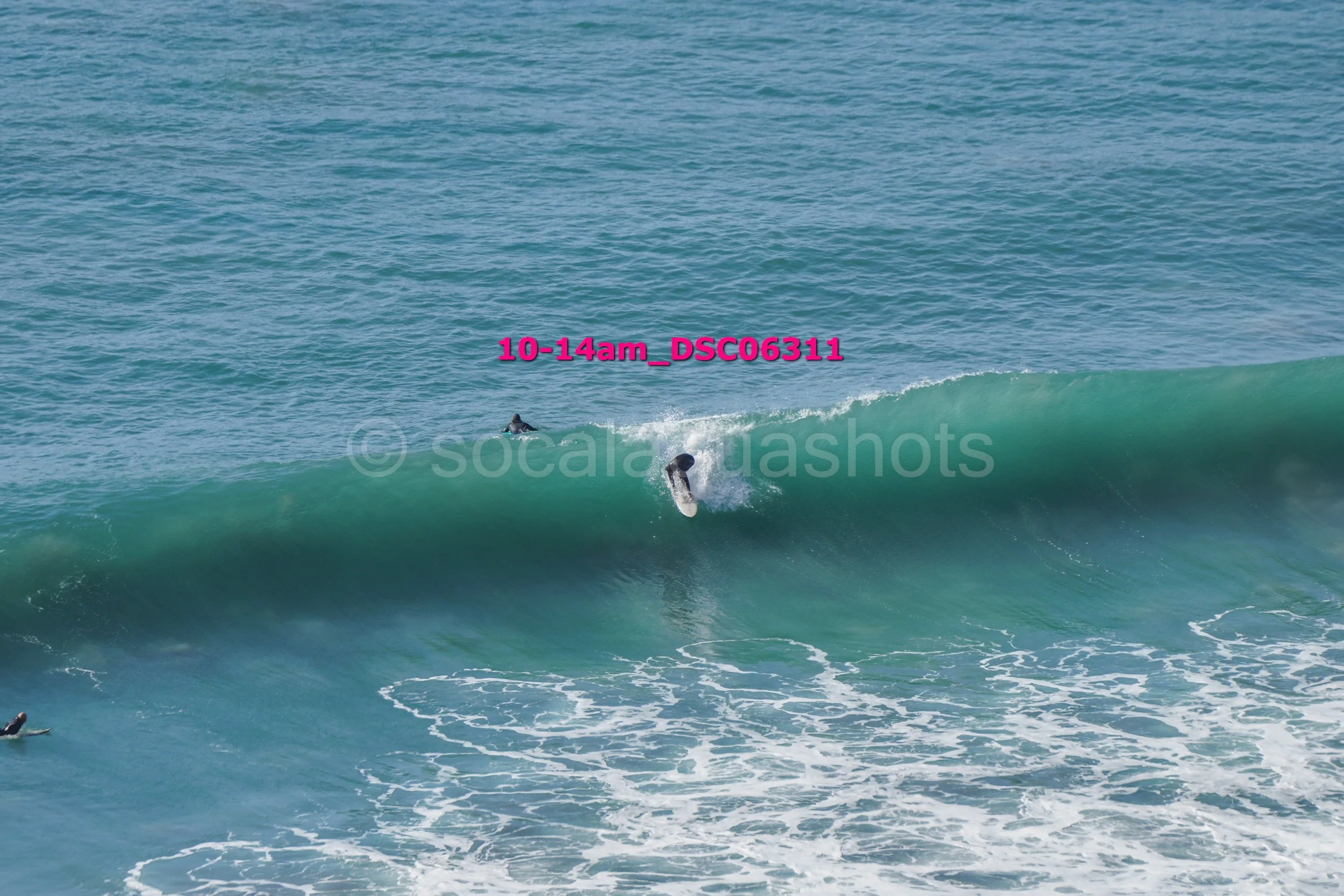 A person surfing on a wave in the ocean with two other surfers in the water. The image has pink text overlay and a watermark.