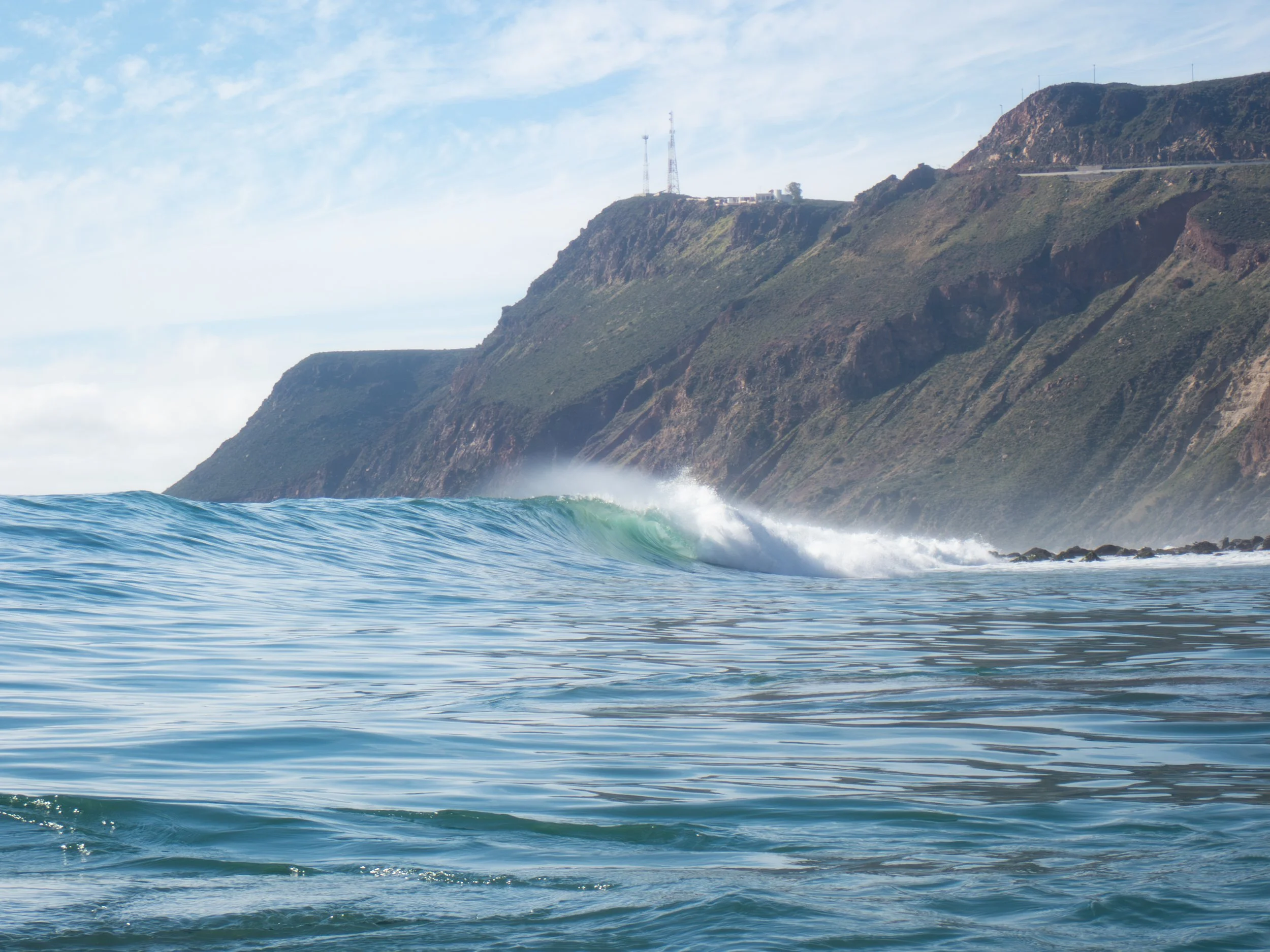 Ocean waves near a rocky shoreline with rugged cliffs and hills in the background under a partly cloudy sky.