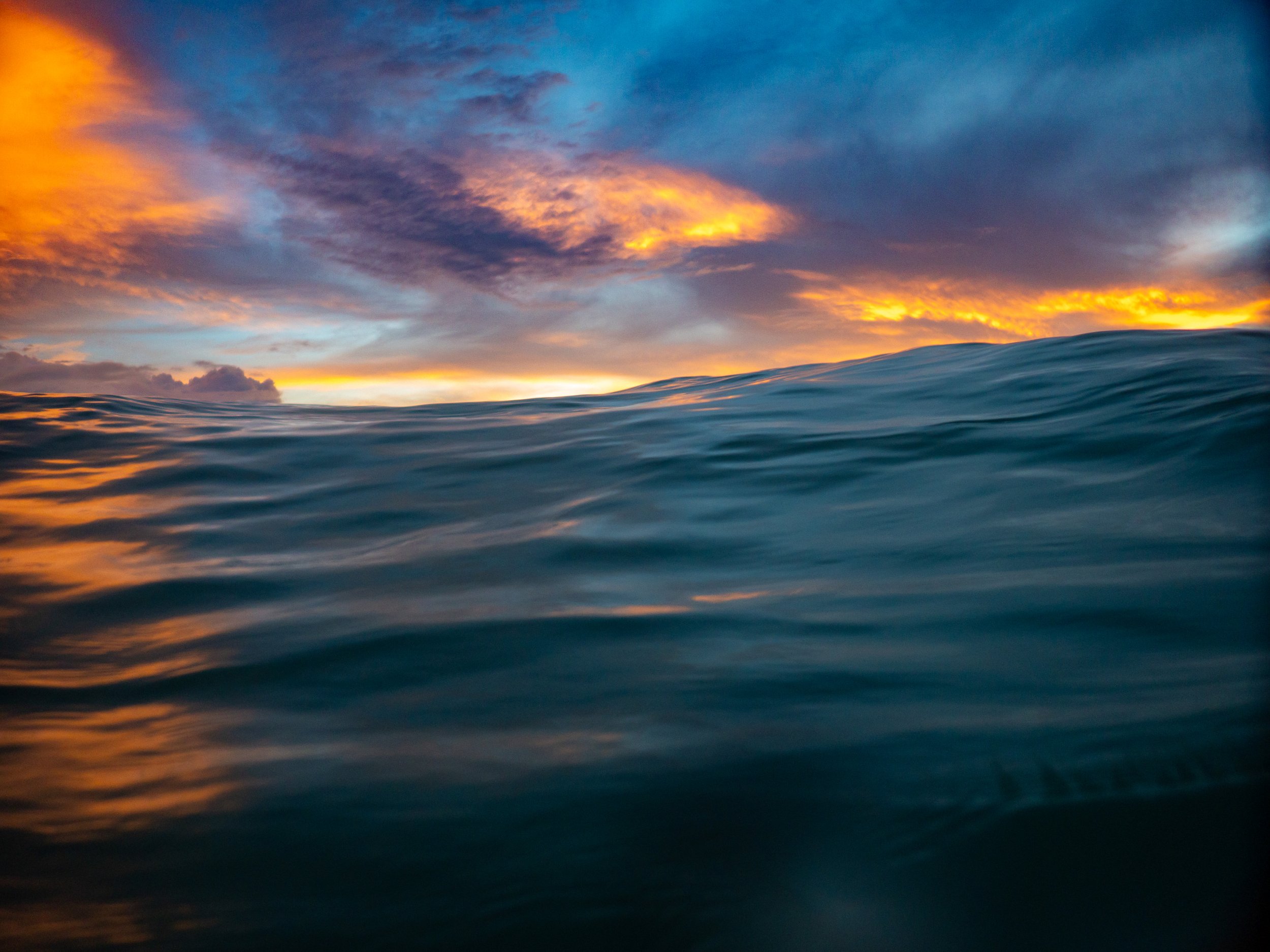 Ocean wave with a colorful sunset sky in the background, displaying shades of orange, yellow, blue, and purple.