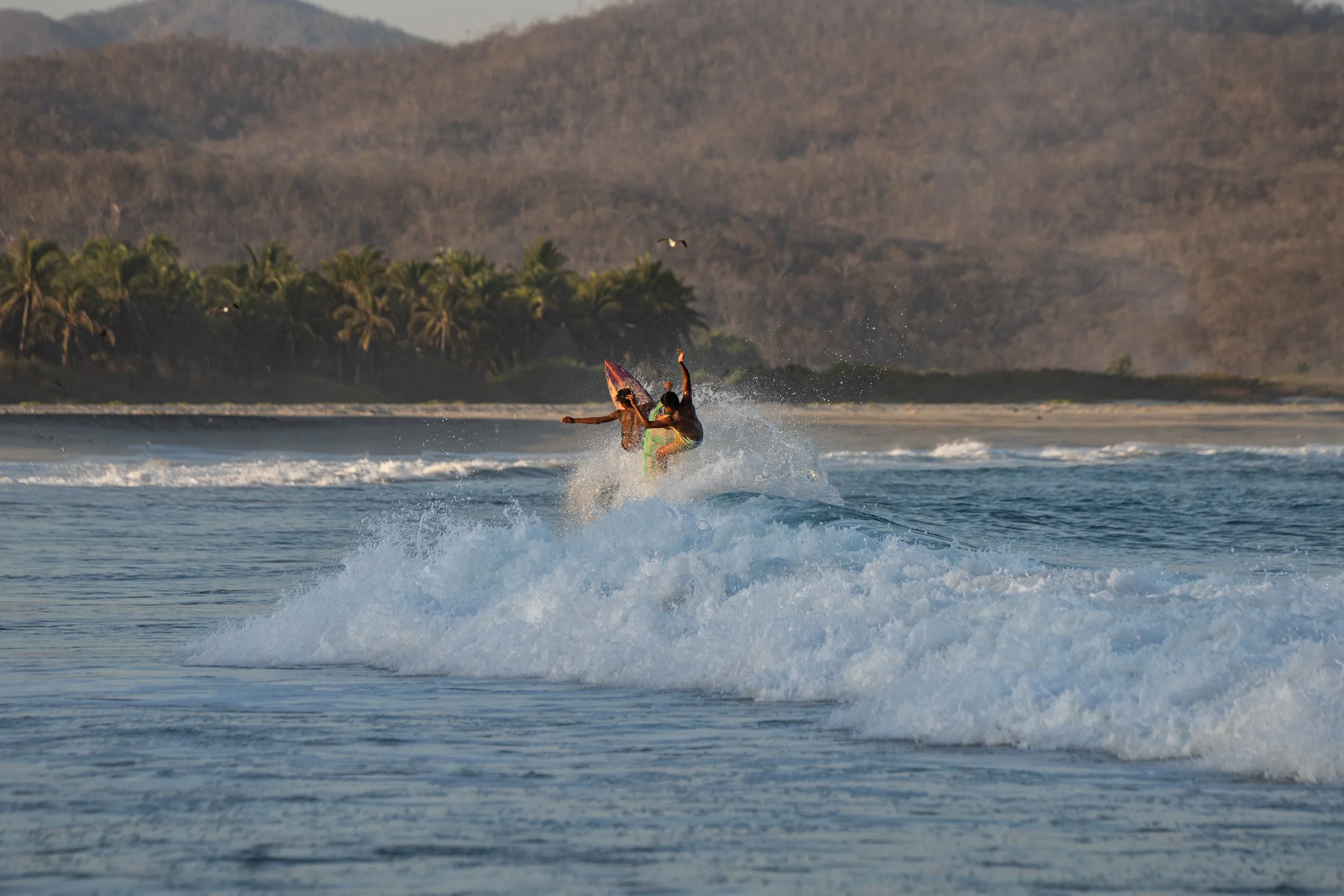 Two people surfing on the ocean with a tropical coastline and mountains in the background.