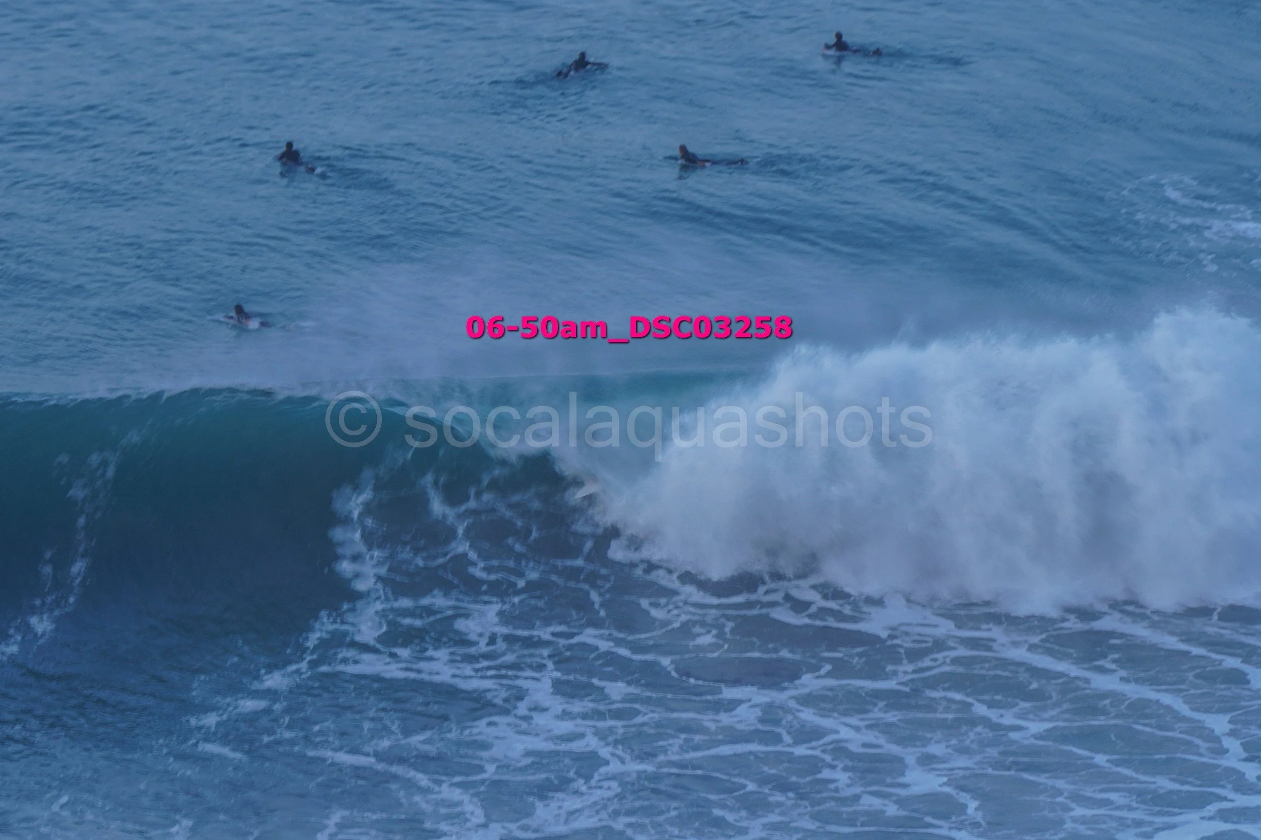 Group of surfers waiting on their surfboards in the ocean as a large breaking wave approaches.