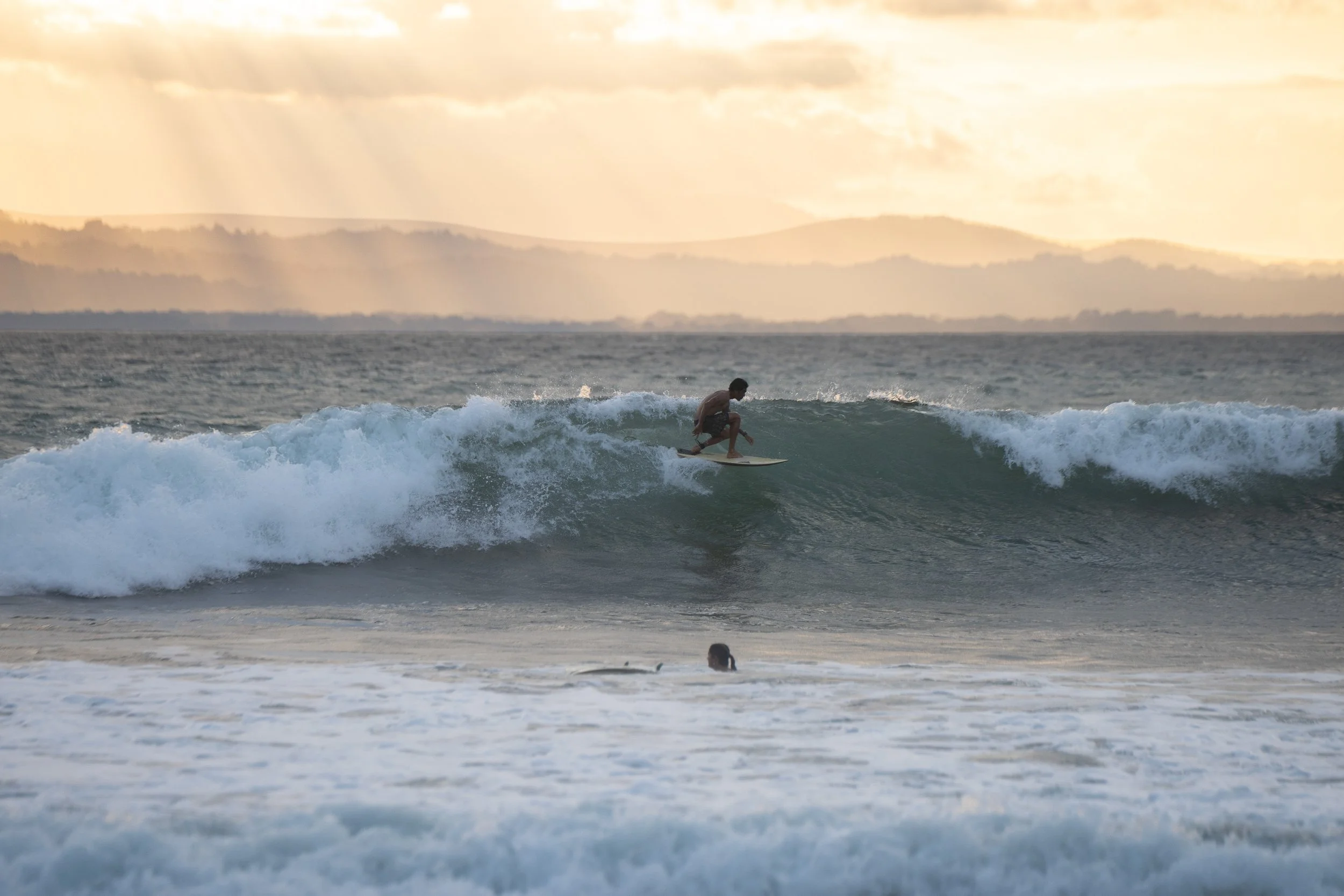 A person surfing on a wave in the ocean during sunset with mountains in the background.