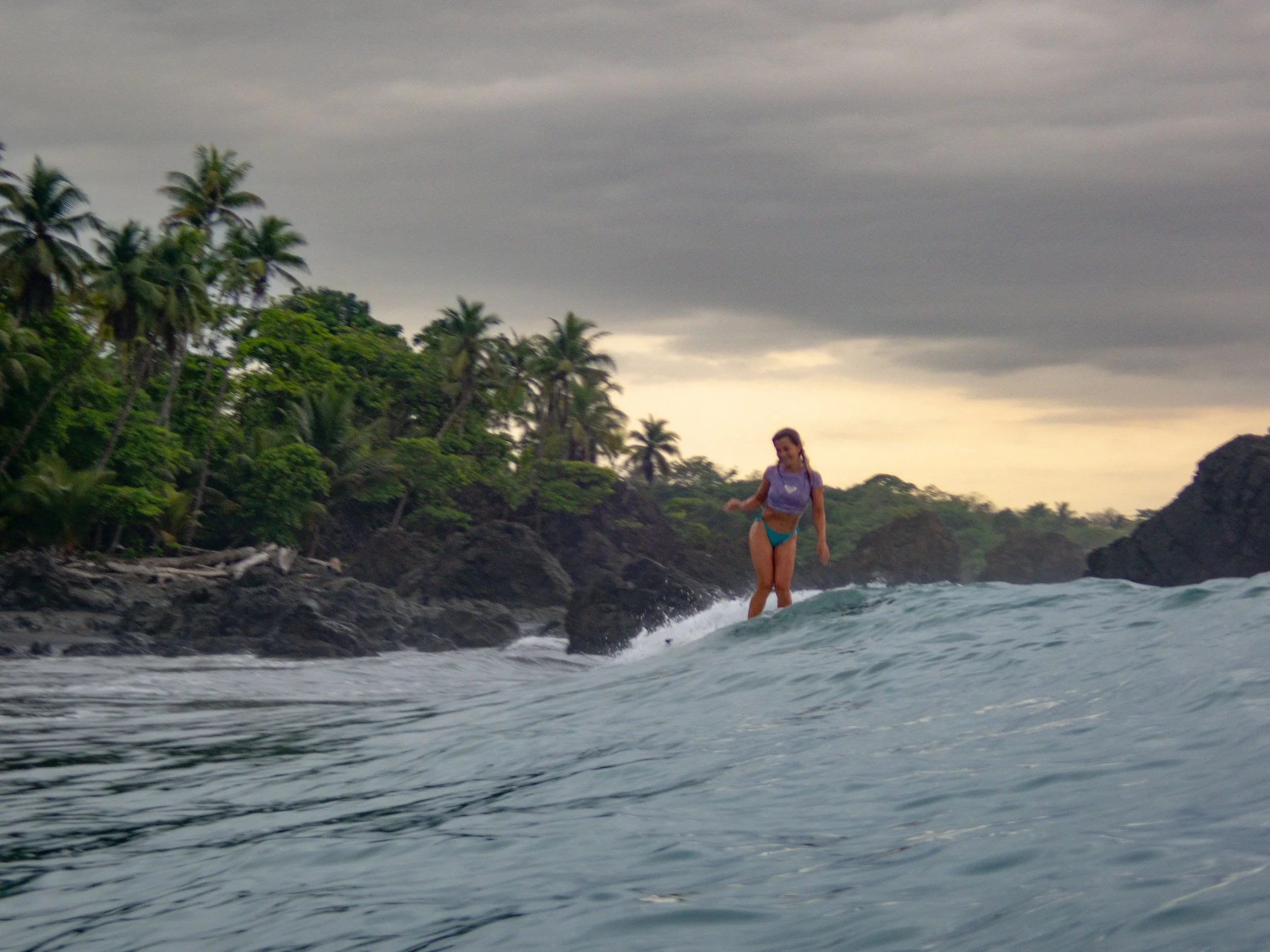Person surfing a wave near a rocky beach with palm trees in the background under an overcast sky.