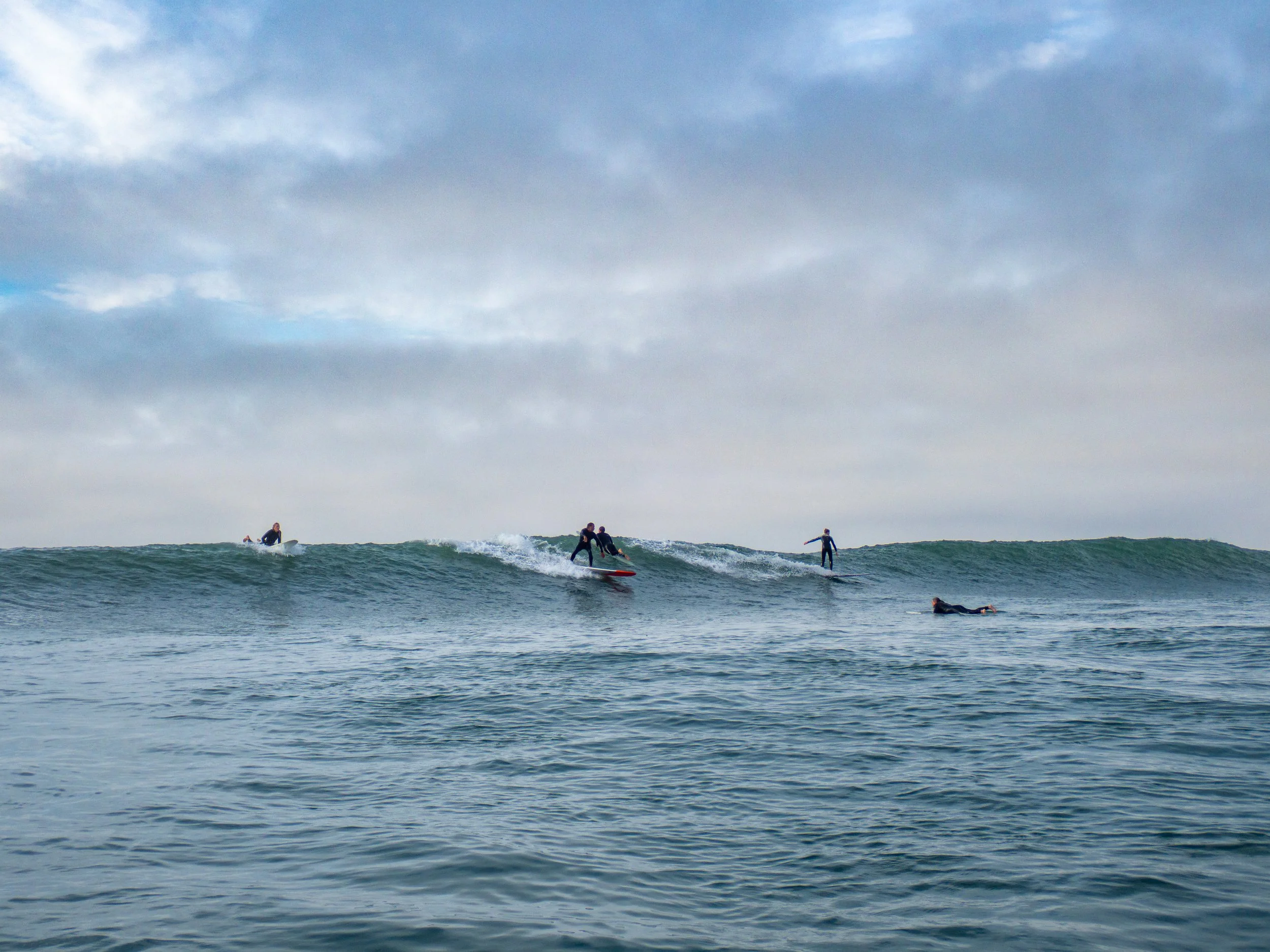 Several surfers riding waves and one swimmer in the water under a cloudy sky.