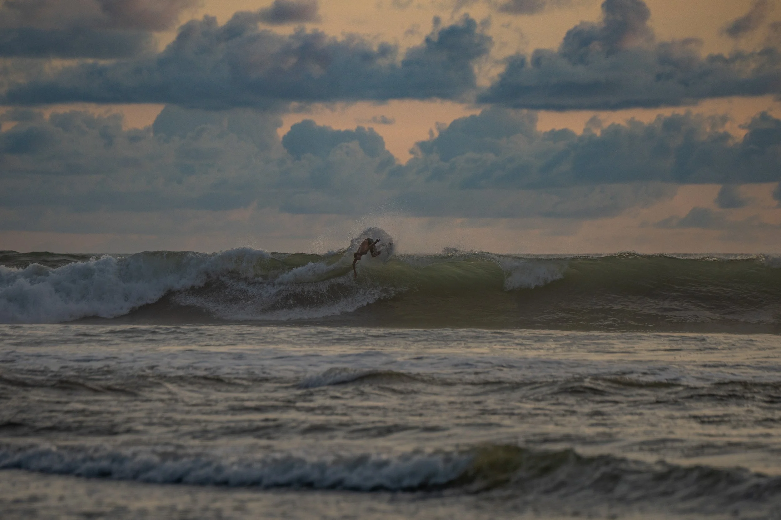 A person surfing a wave in the ocean during dusk or dawn, with cloudy sky overhead.