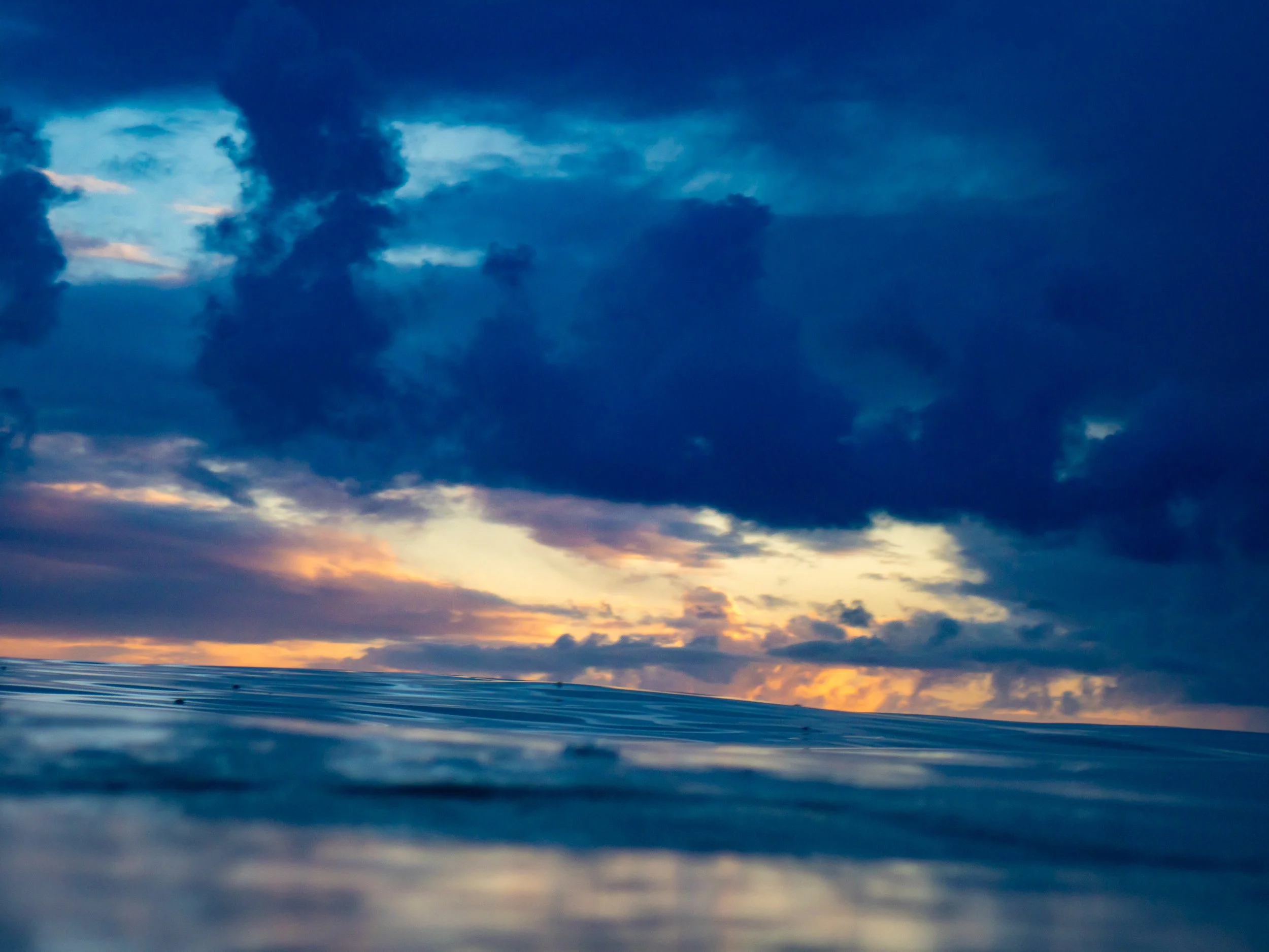 A view of the ocean with a partly cloudy sky at sunset, featuring dark clouds and a hint of orange and yellow light near the horizon.