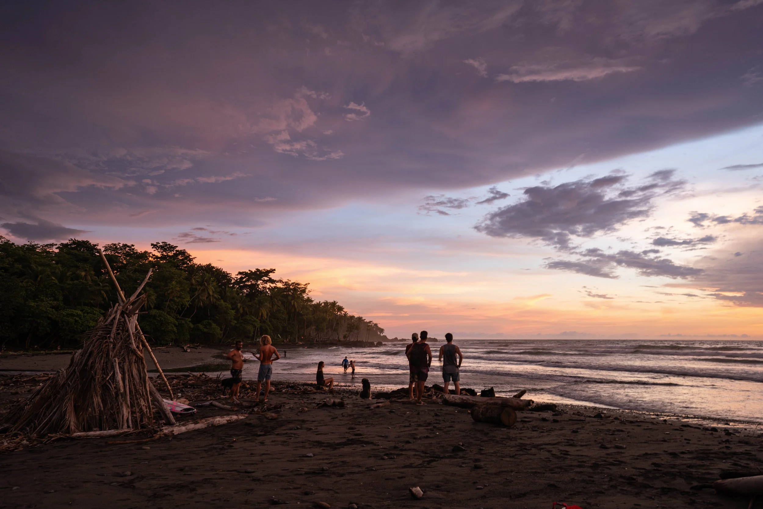 People walking and socializing on a dark sandy beach during sunset, with a cloudy sky and lush tropical trees in the background.