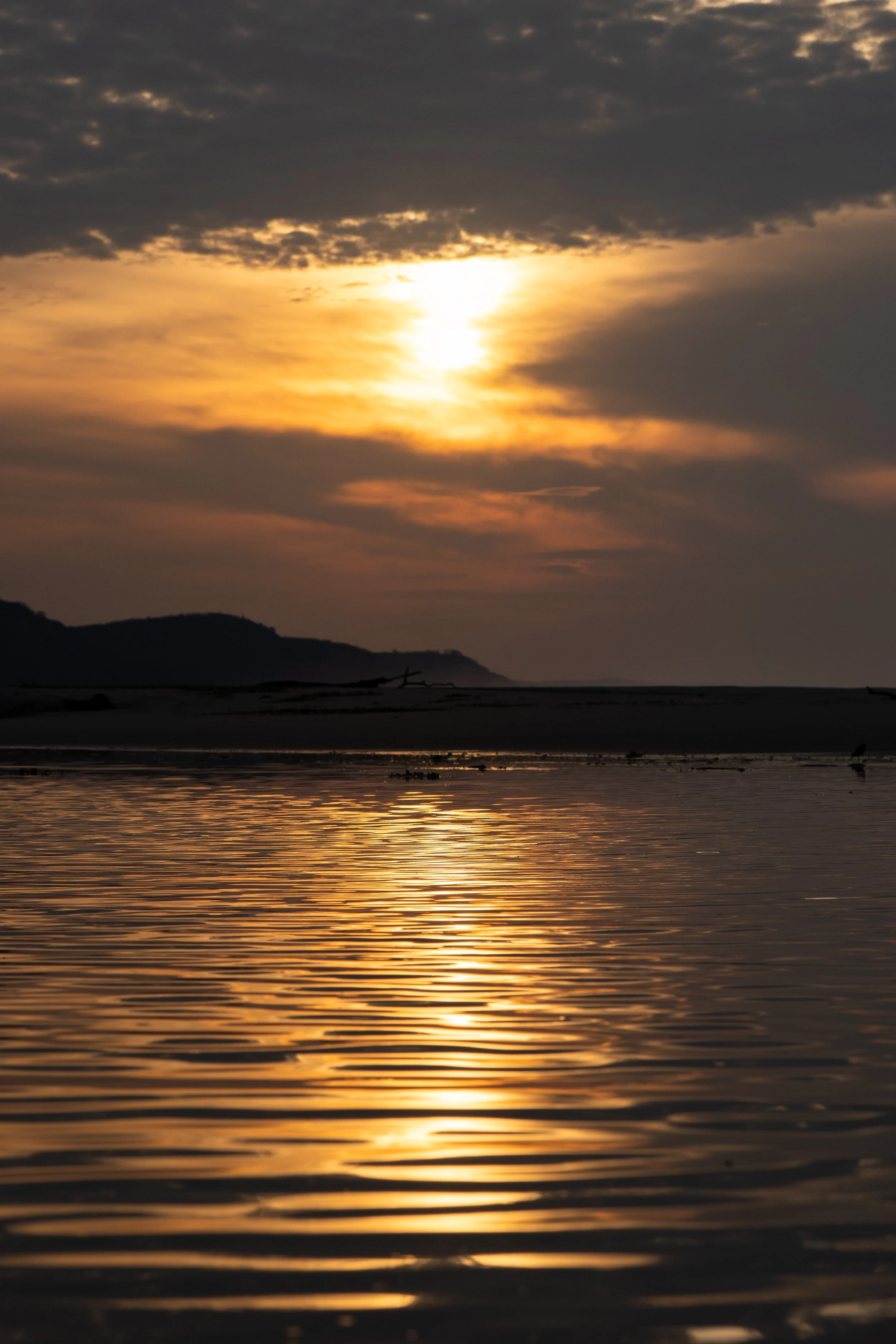 A sunset over a body of water with clouds in the sky and a silhouette of distant hills.