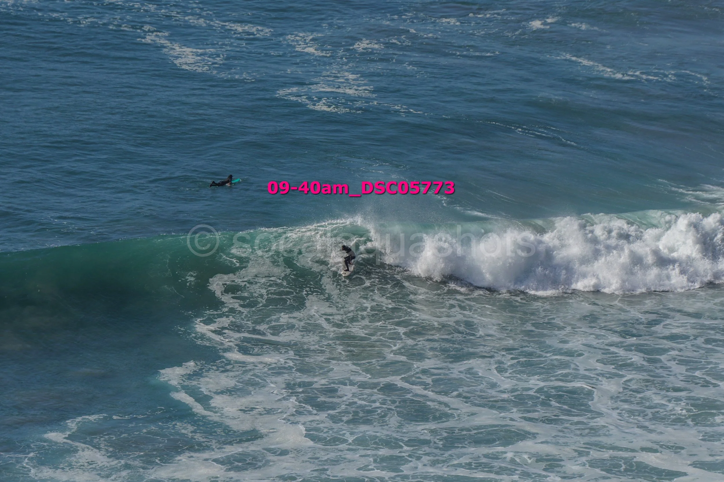 A person surfing on a wave in the ocean, with another person floating on a surfboard nearby.