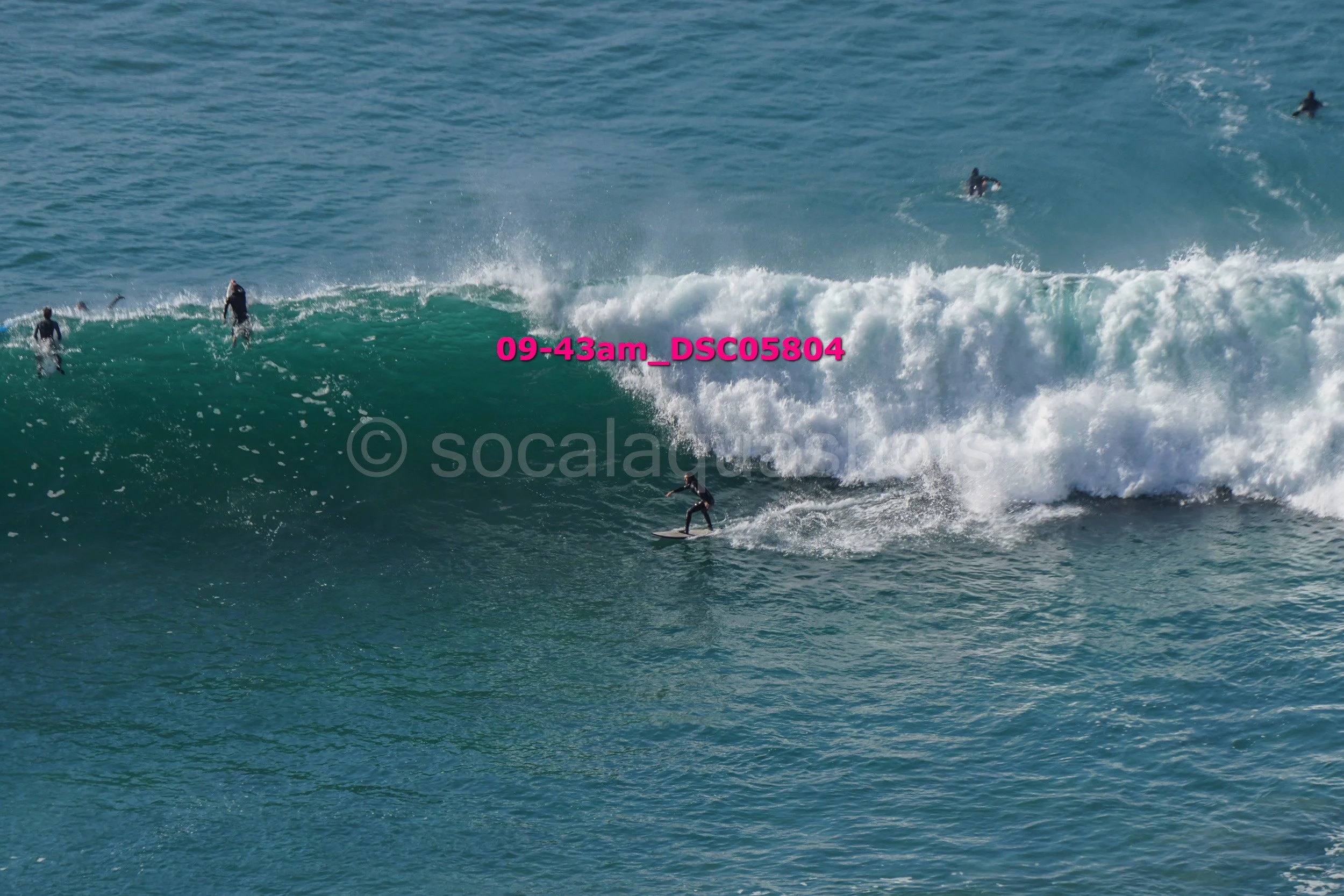 A surfer riding a large wave in the ocean with multiple other surfers visible in the water around the wave.