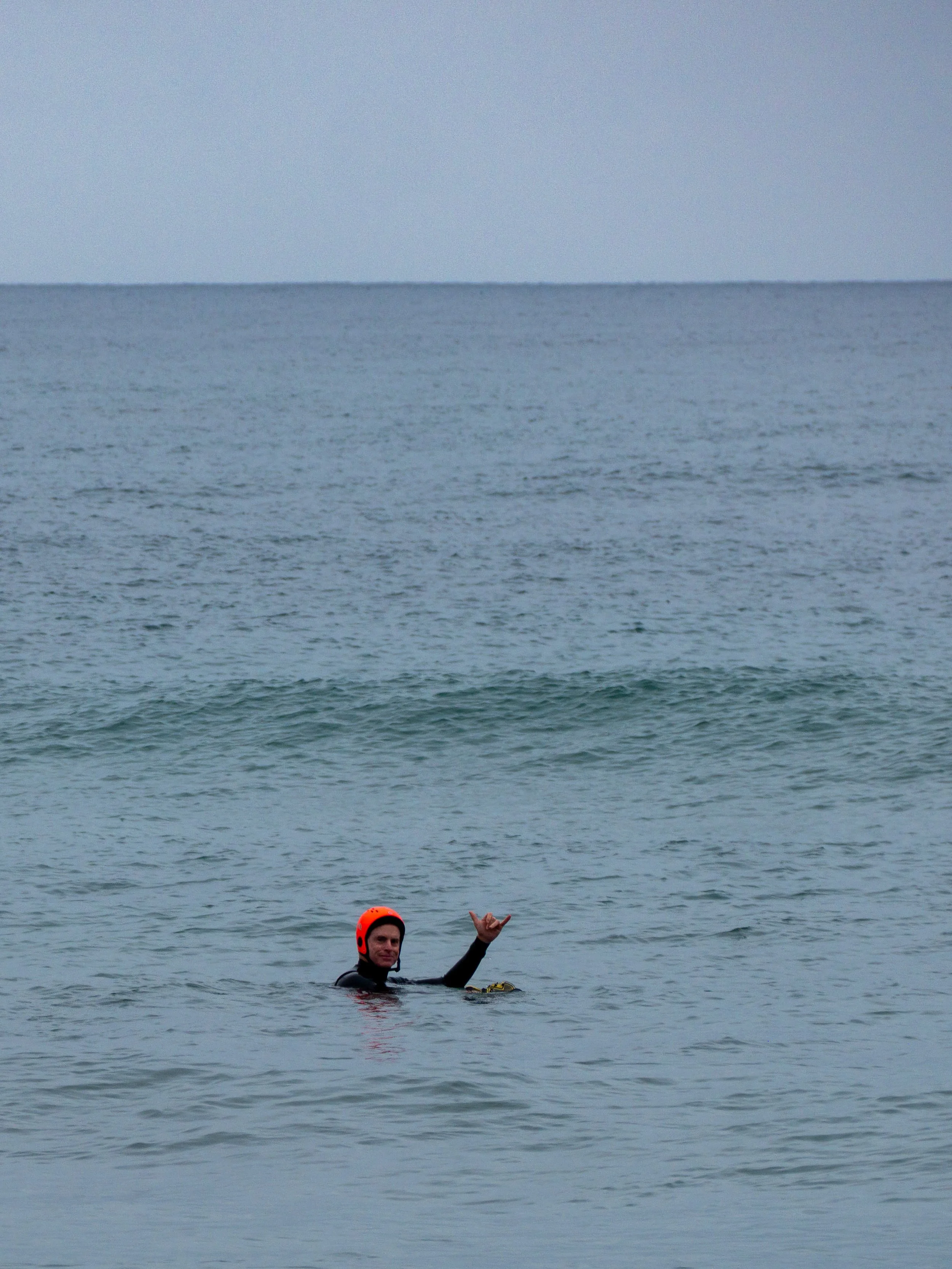 A person in a wetsuit and orange helmet swimming in the ocean, making a shaka sign with their hand.