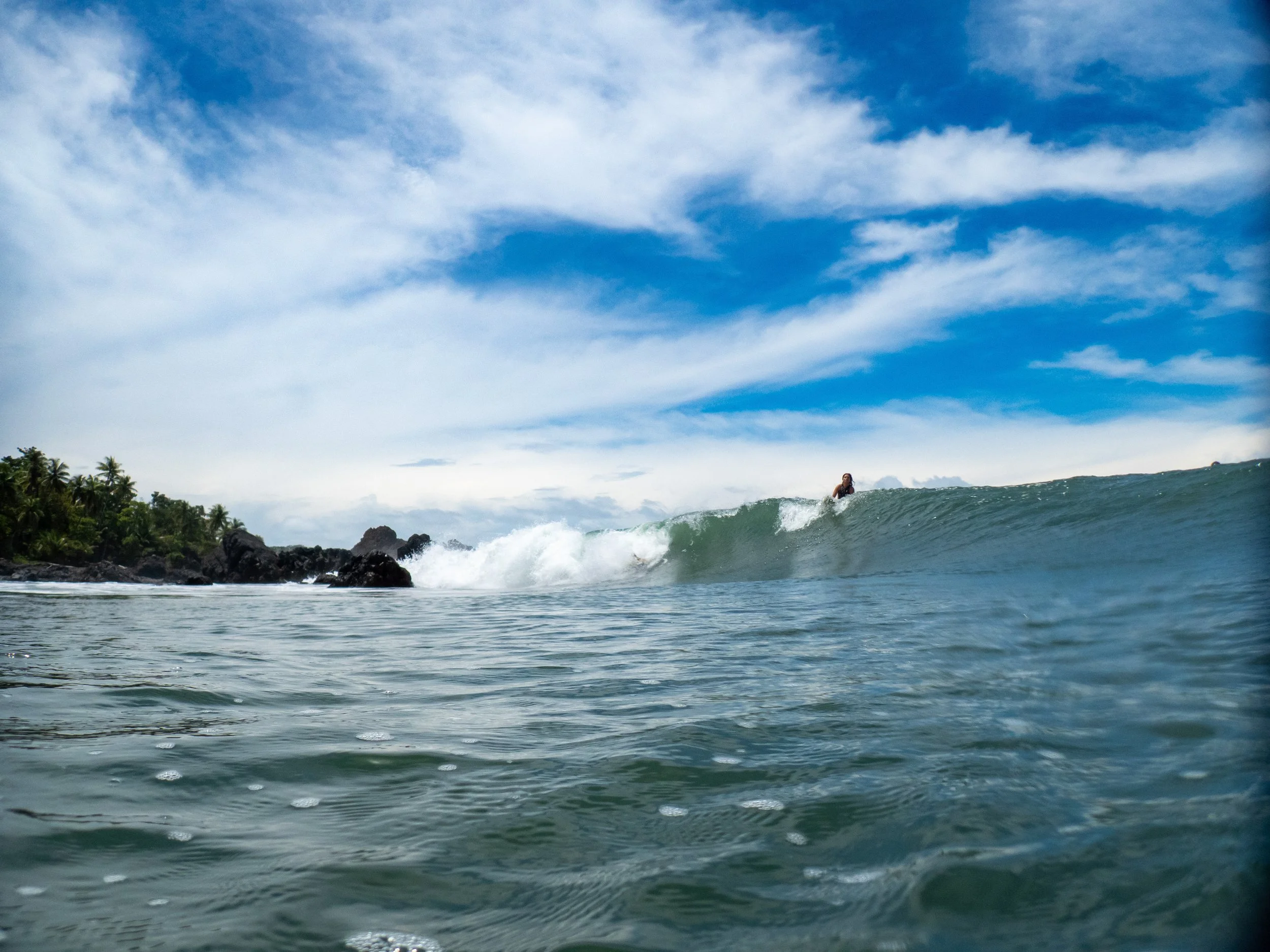 Ocean with a surfer riding a wave near a tropical island with palm trees and rocky shoreline, under a partly cloudy sky.