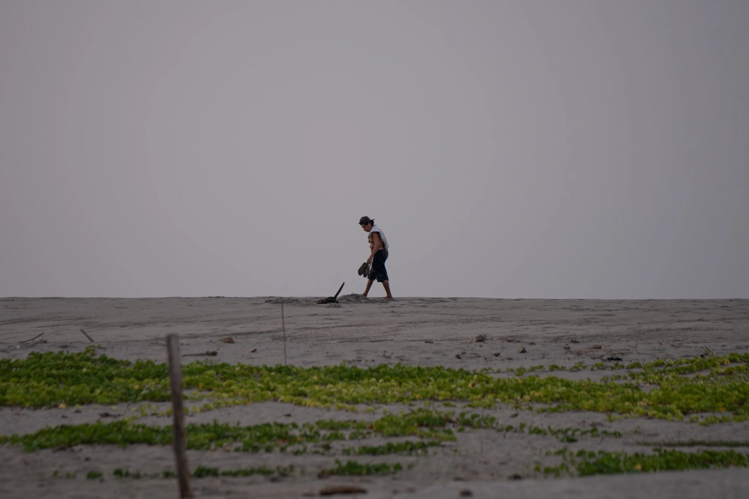 A person walking on a sandy beach carrying a skateboard with a gray sky in the background.