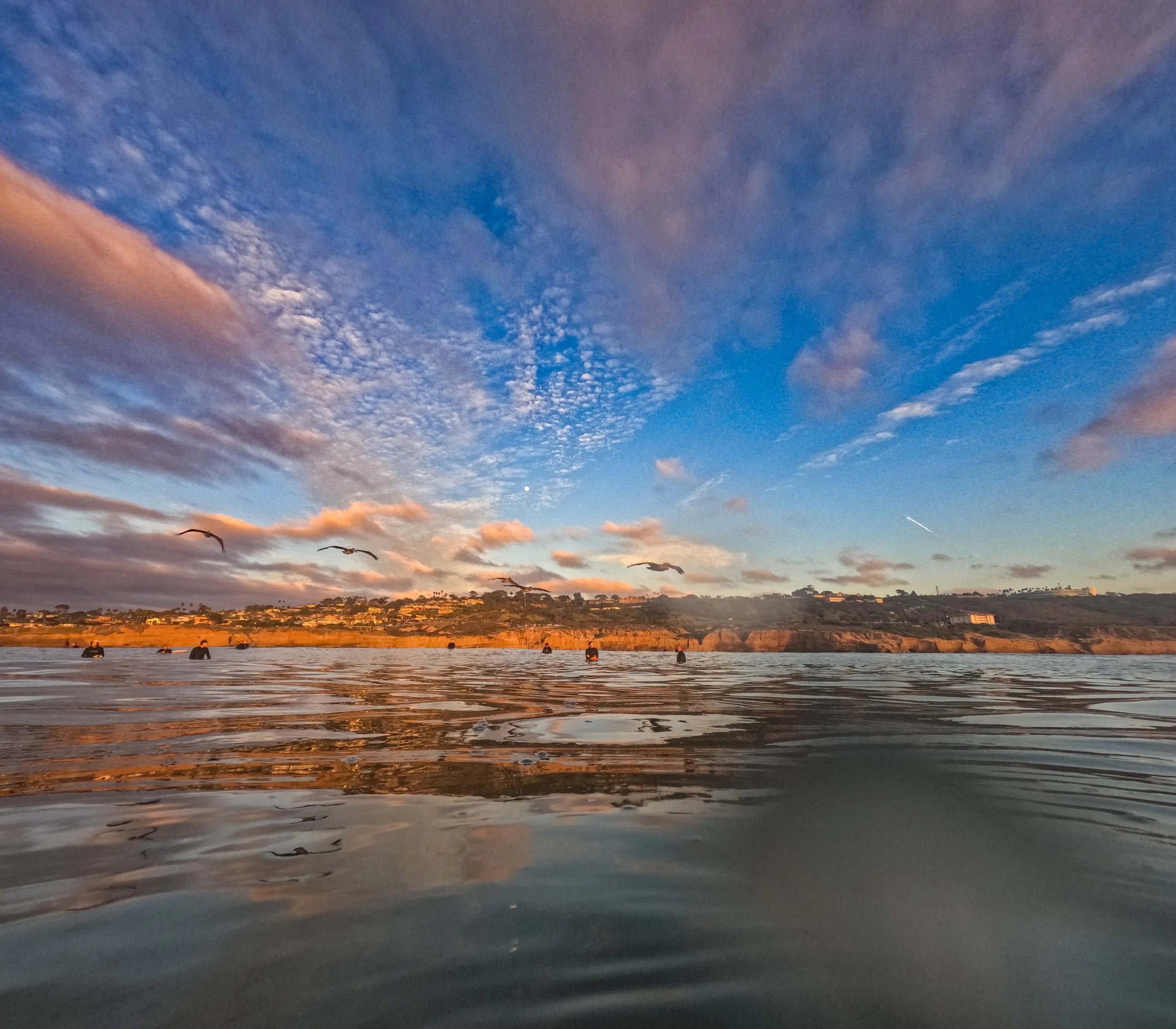 Sunset over the ocean with birds flying and some boats floating on the water, and a distant hillside with buildings against a sky with clouds.