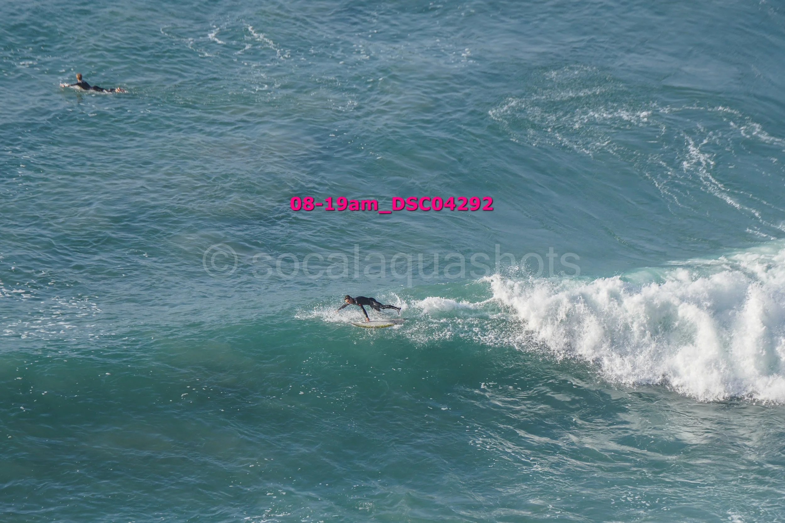 Surfer riding a wave in the ocean with another surfer swimming nearby.