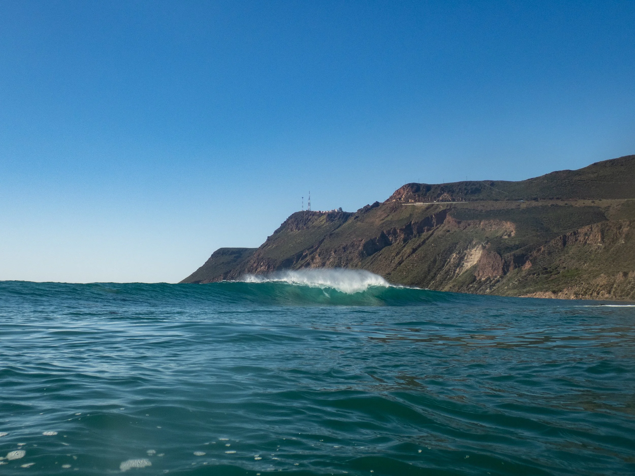 The image shows an ocean with a wave in the foreground and a rugged coastline with hills in the background under a clear blue sky.