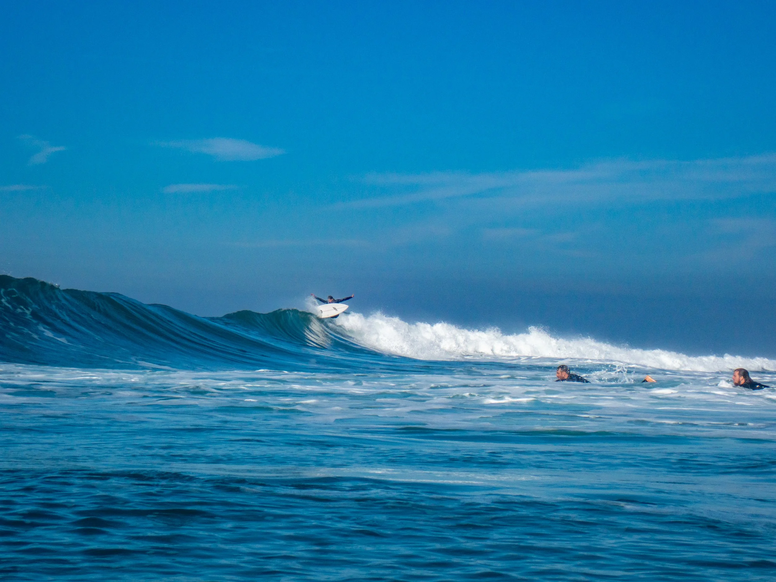 Surfer riding a wave on a clear day, with two other surfers in the water watching, under a blue sky with some clouds.