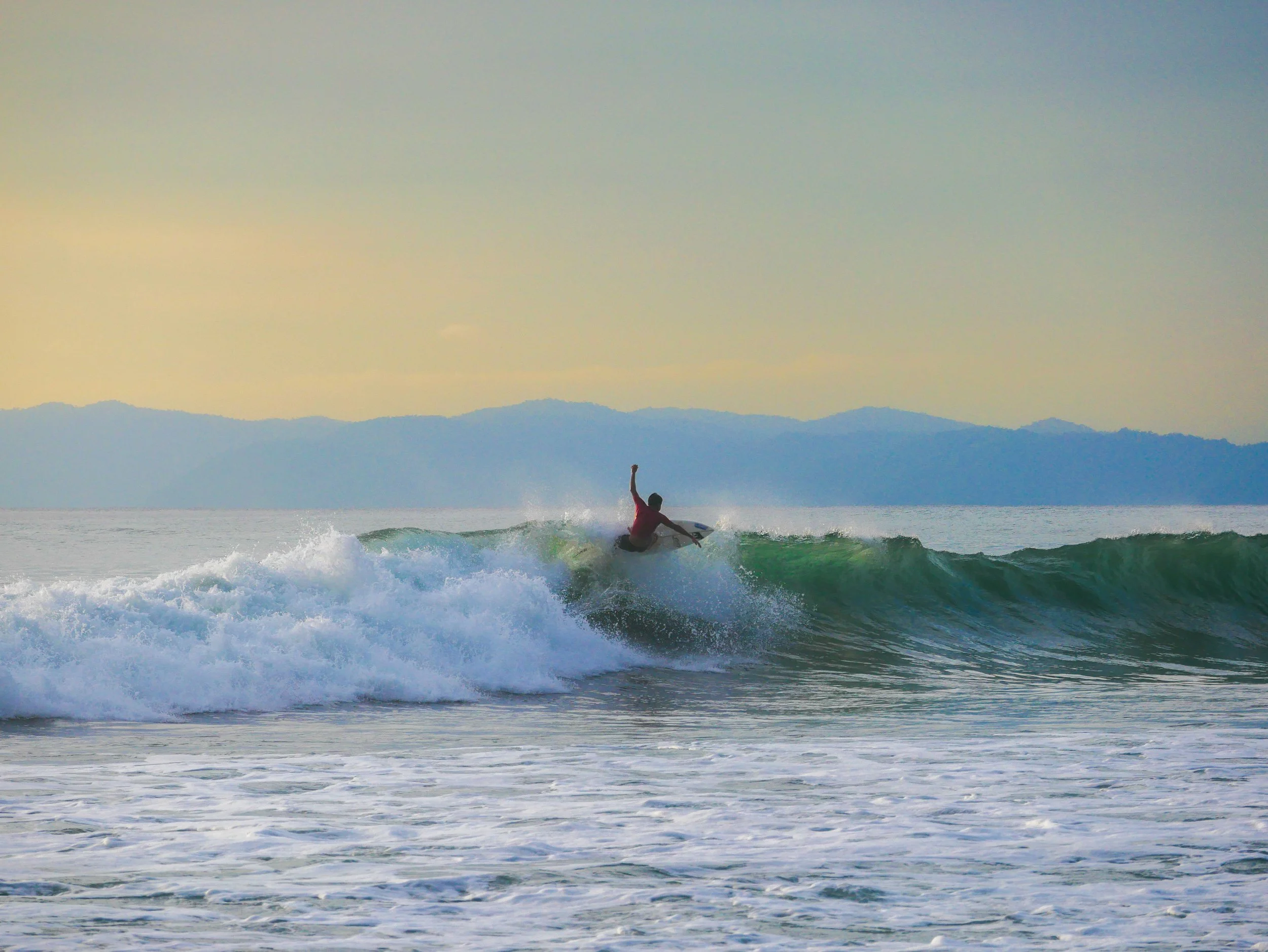 Surfer riding a wave at sunset with mountains in the background.
