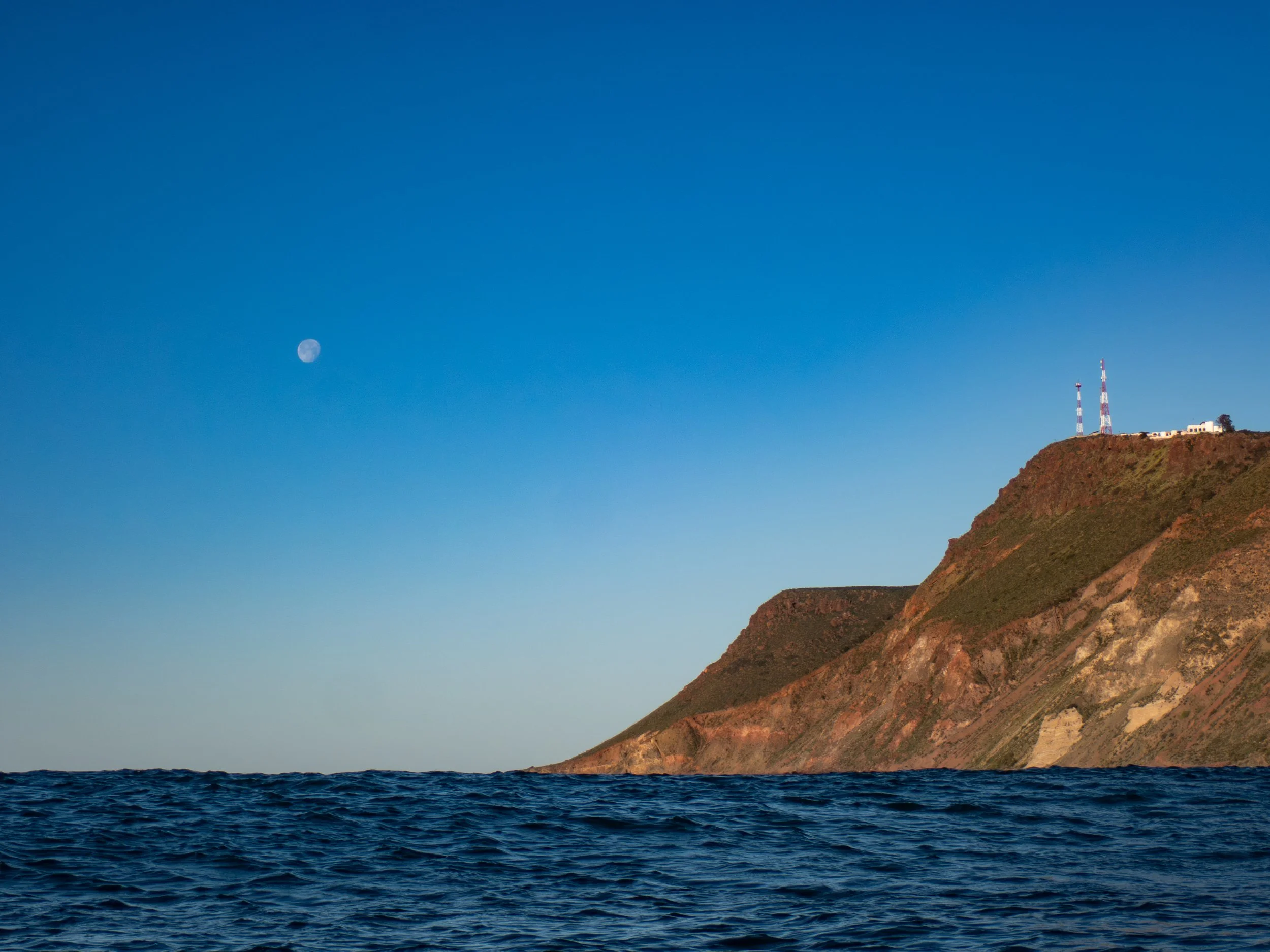 Ocean with dark blue waves, coastline with steep hills, and antennas on top of the hill, under a clear sky with visible moon.
