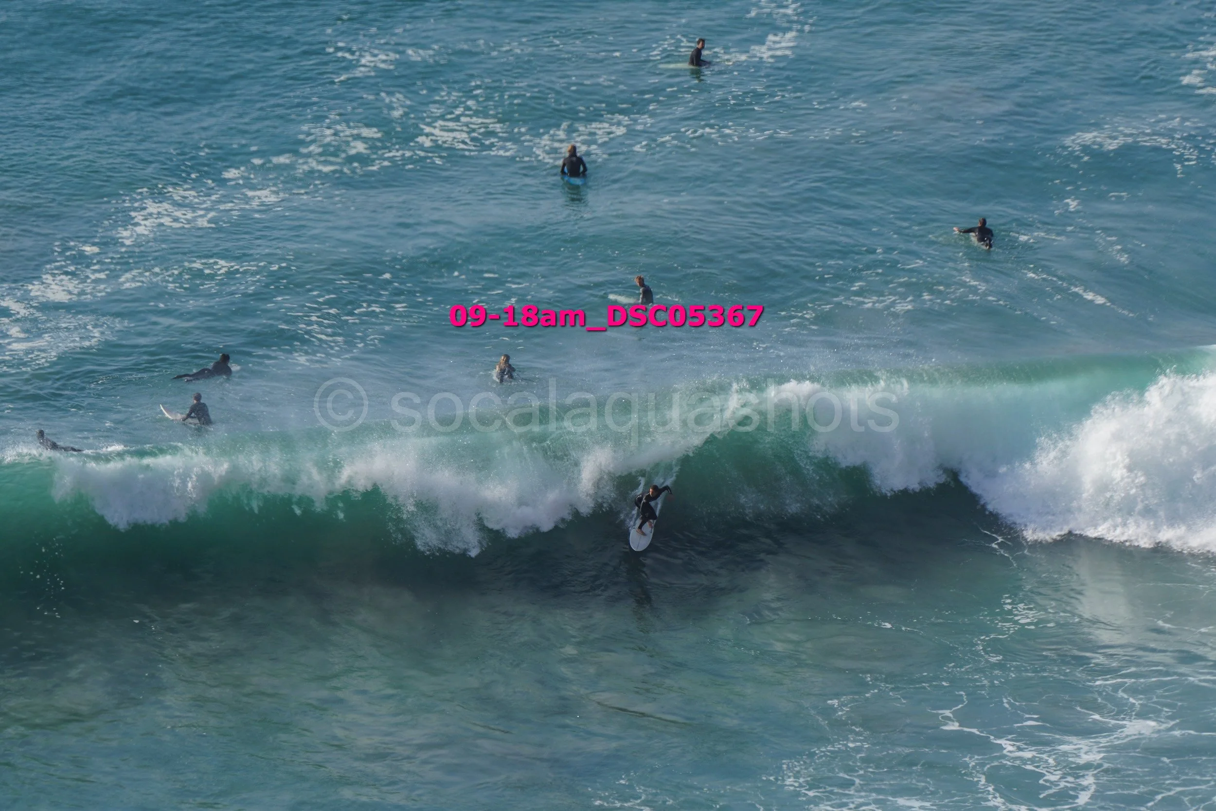 Surfer riding a wave with several other surfers in the ocean, visible in the background.