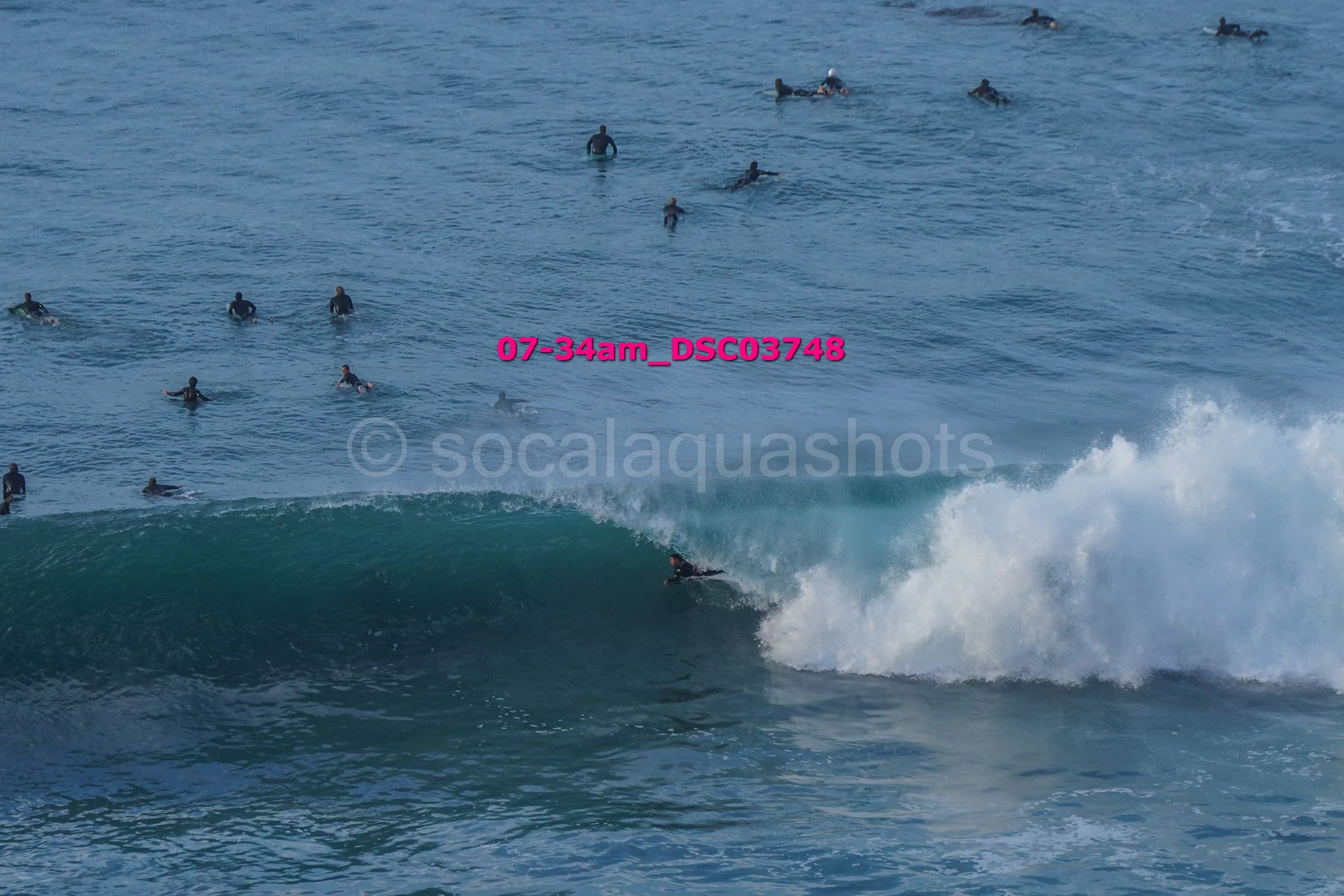 Surfer riding a wave in the ocean with many people swimming and floating in the water nearby.
