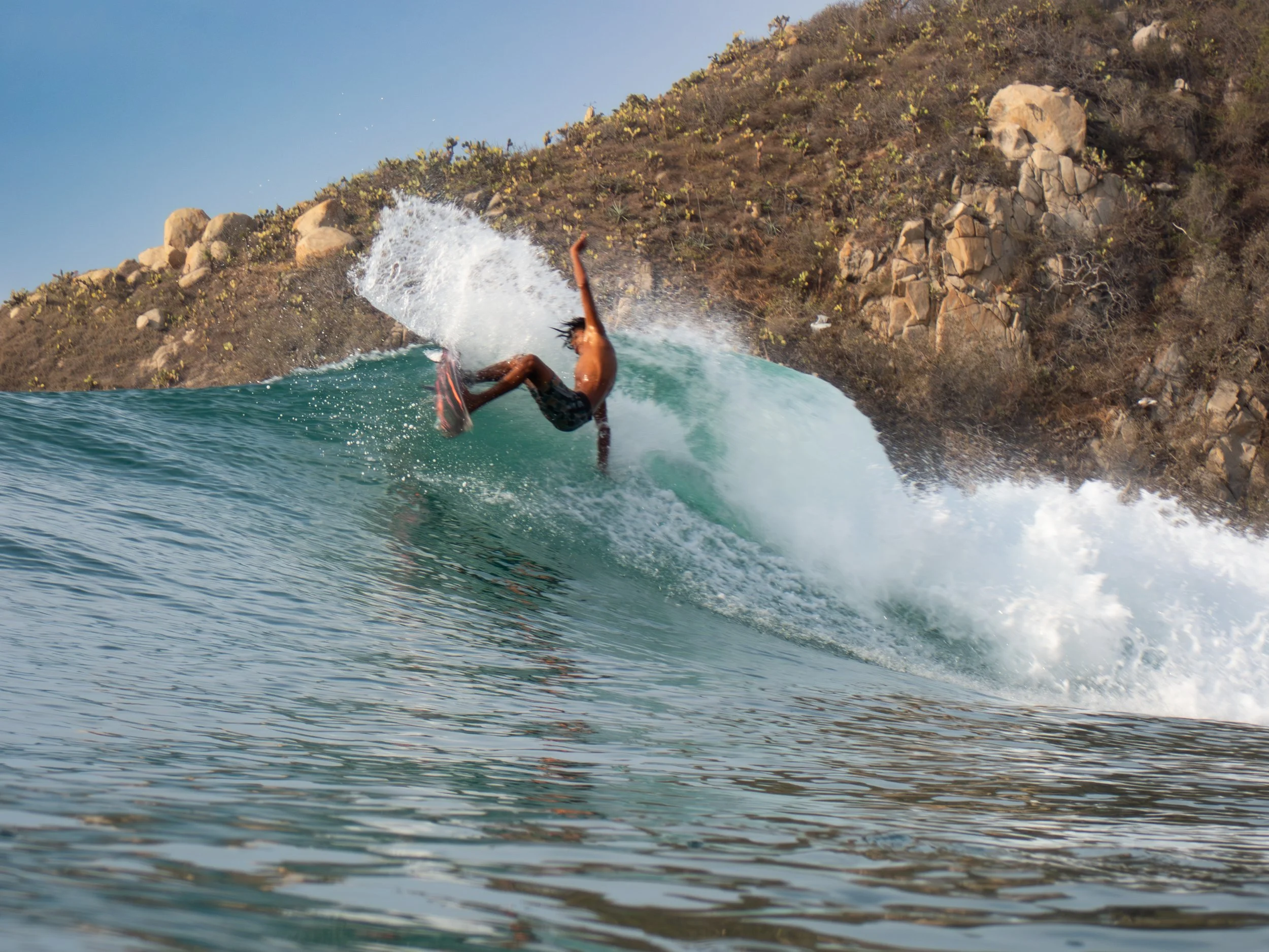 A person surfing on a wave near a rocky hillside with sparse desert vegetation.