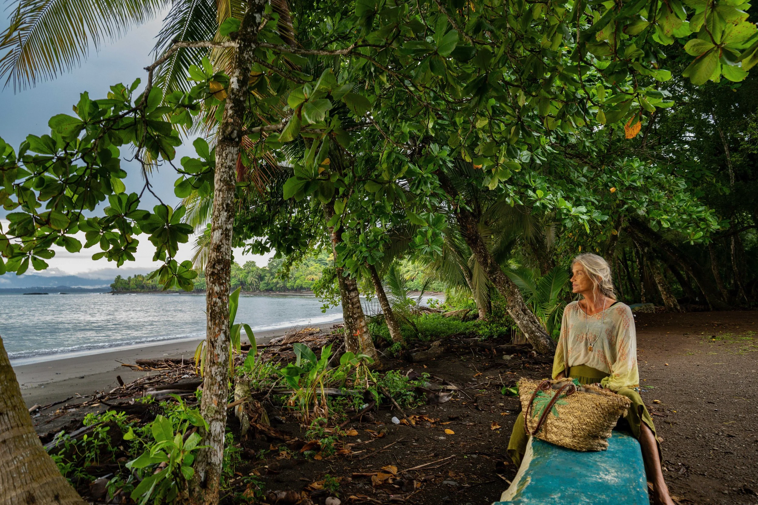 A woman with long gray hair sitting on a beach after sunset, surrounded by lush green tropical trees and foliage, with a large woven bag on her lap, looking to the side.