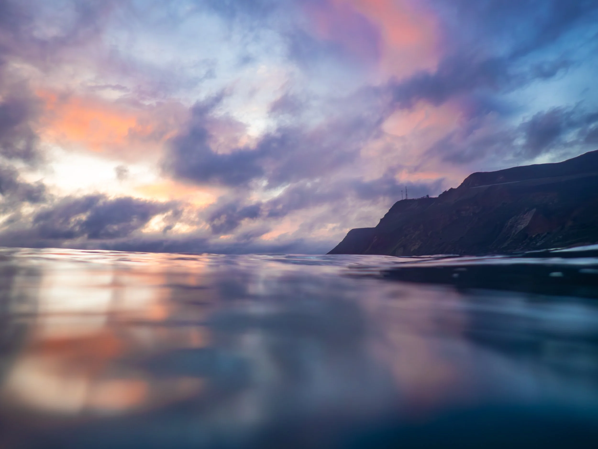 Scenic view of a calm ocean with a rugged mountain in the distance under a colorful sky at dusk.