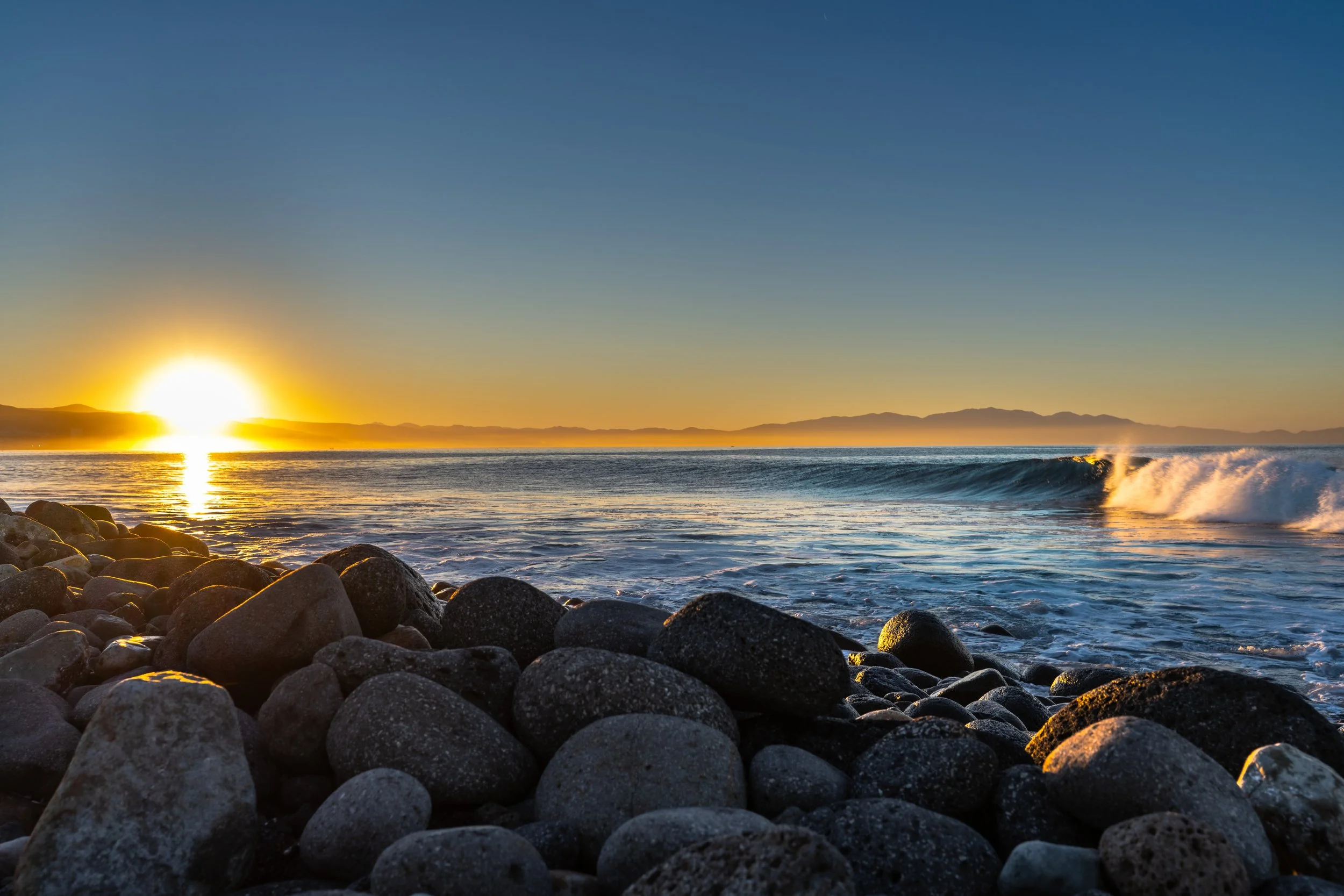 Sunset over the ocean with waves crashing on a rocky beach and distant mountains on the horizon.