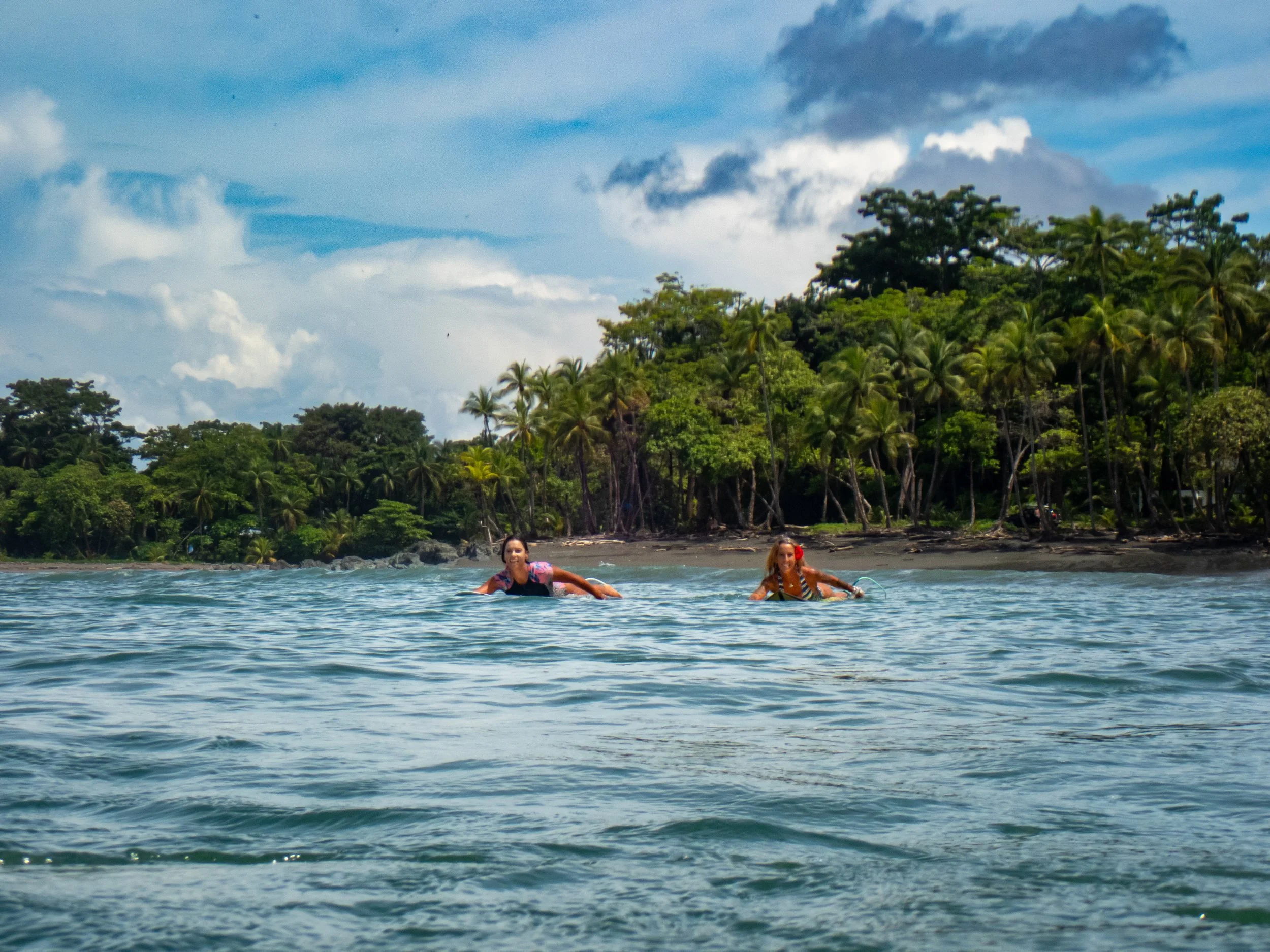 Two women are swimming in a tropical ocean near an island with lush green trees and palm trees, under a partly cloudy sky.