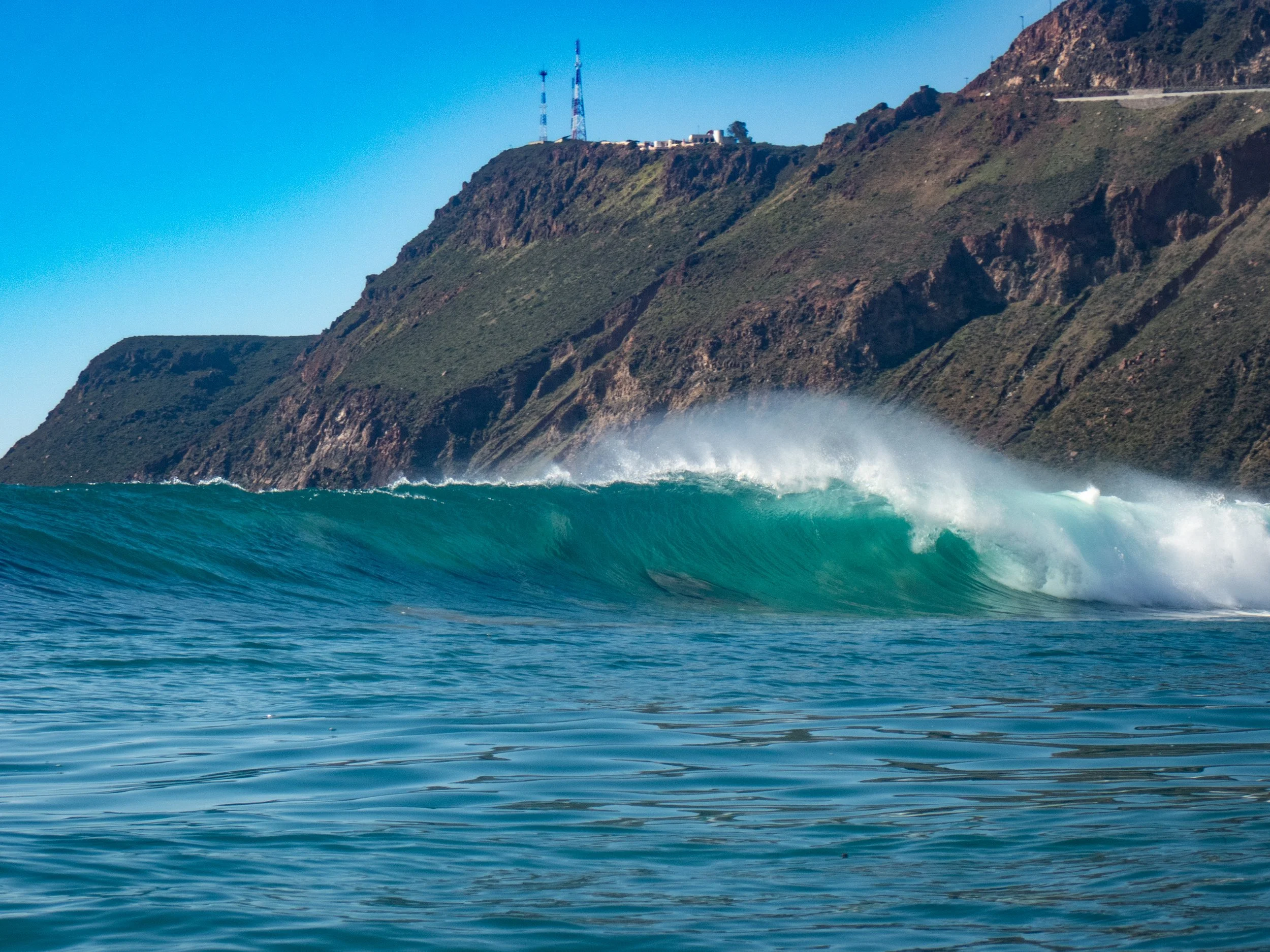 Ocean wave crashing near a rugged green hillside with communication towers on top under a clear blue sky.