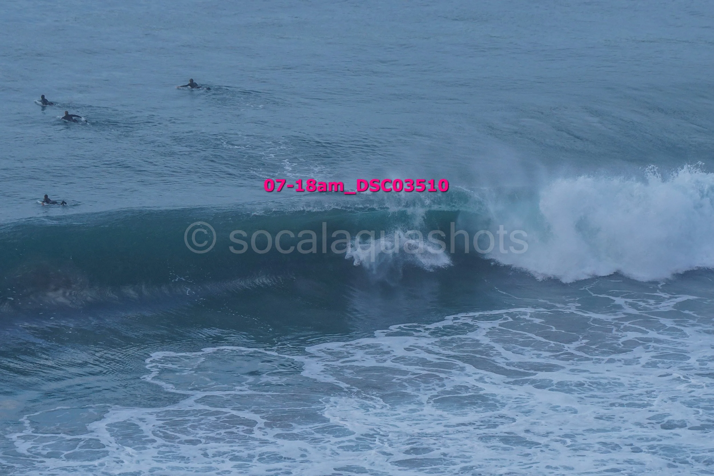 A surf wave with a person riding inside the wave and several other surfers paddling further out in the ocean.