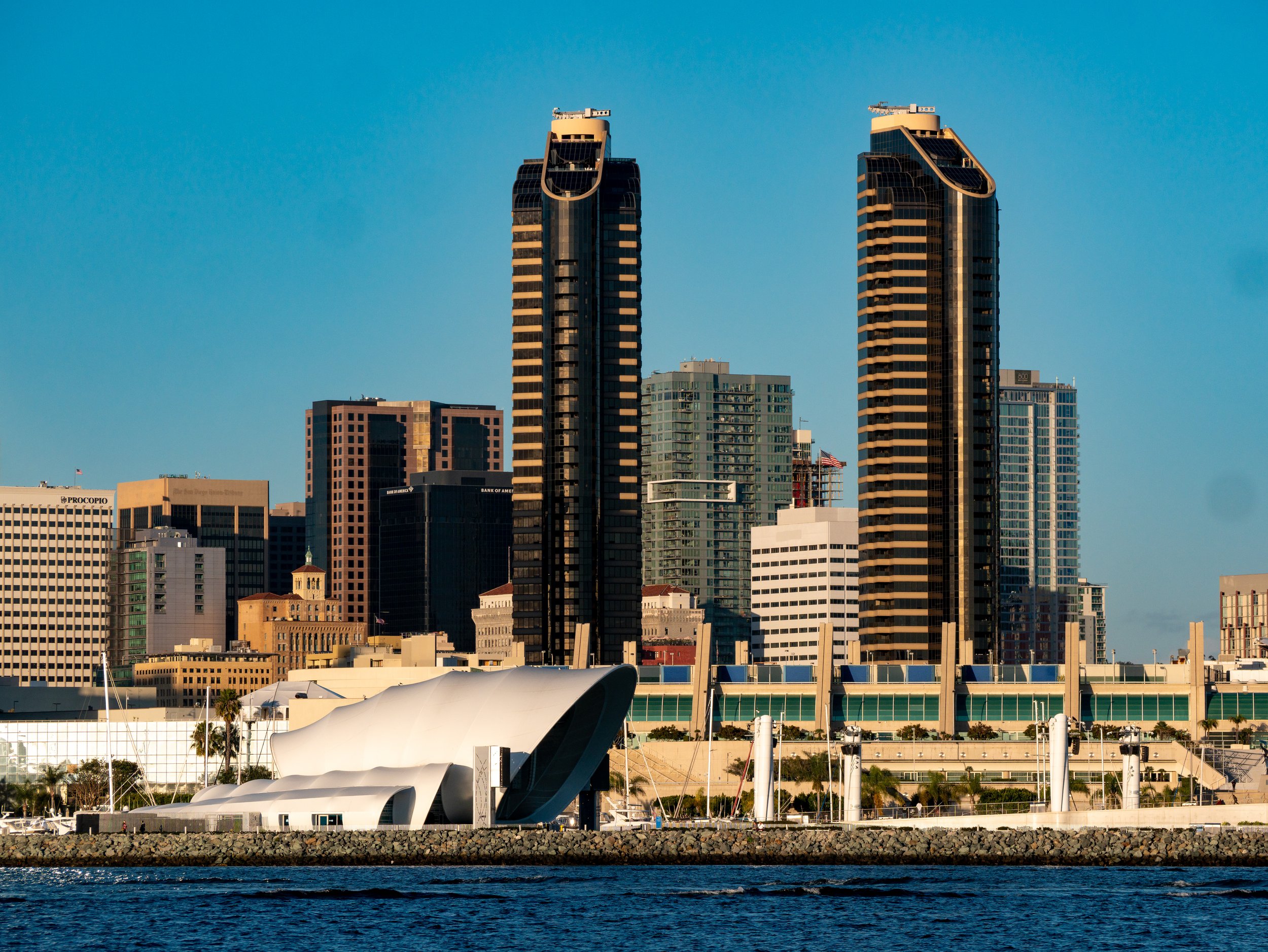 View of a city skyline with tall modern skyscrapers, a white architecturally unique building in the foreground, and a body of water at the bottom.