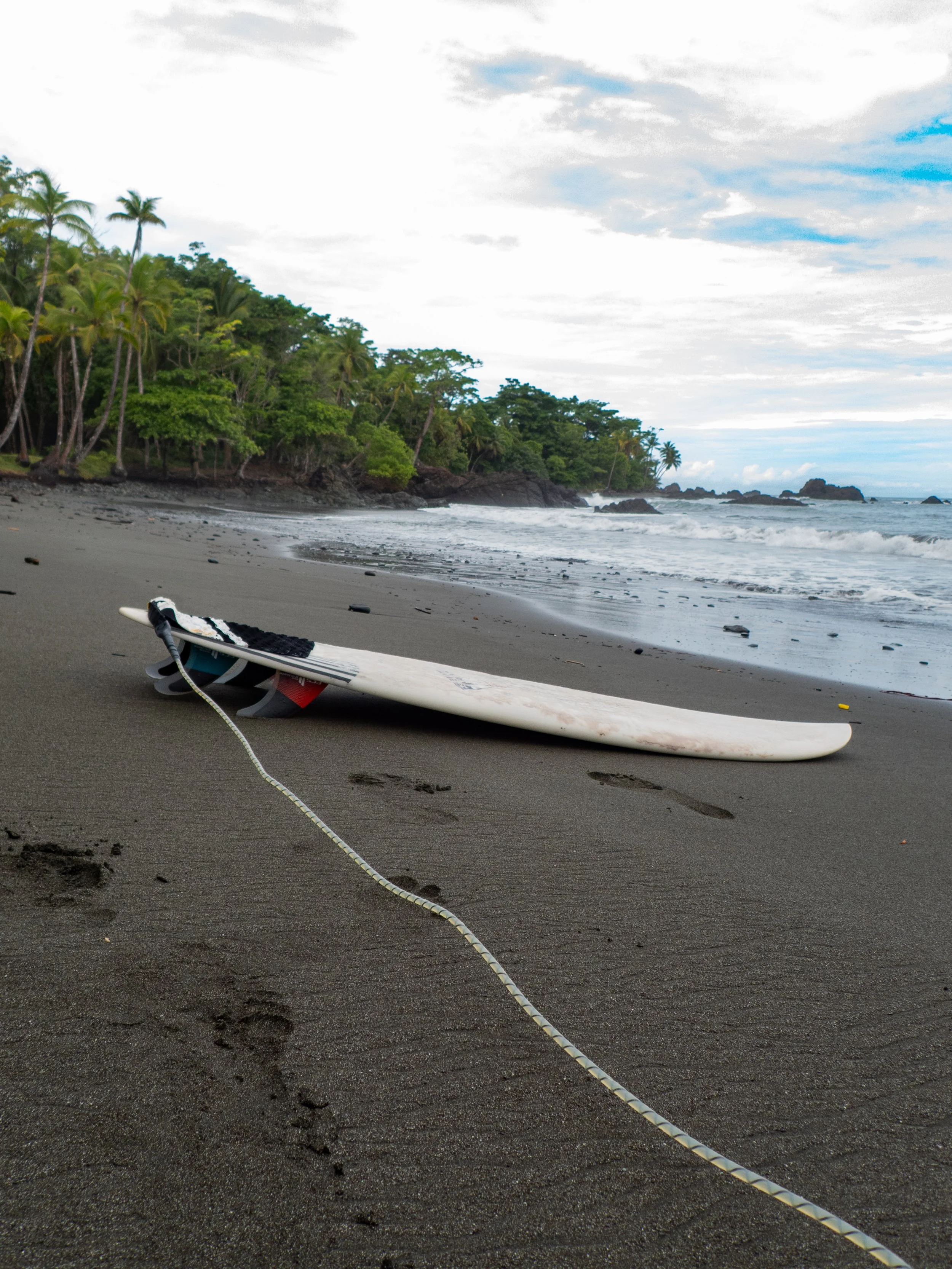 A black and white surfboard lying on dark sandy beach with a rope attached, near footprints, with green trees and rocky shoreline in the background, under cloudy sky.