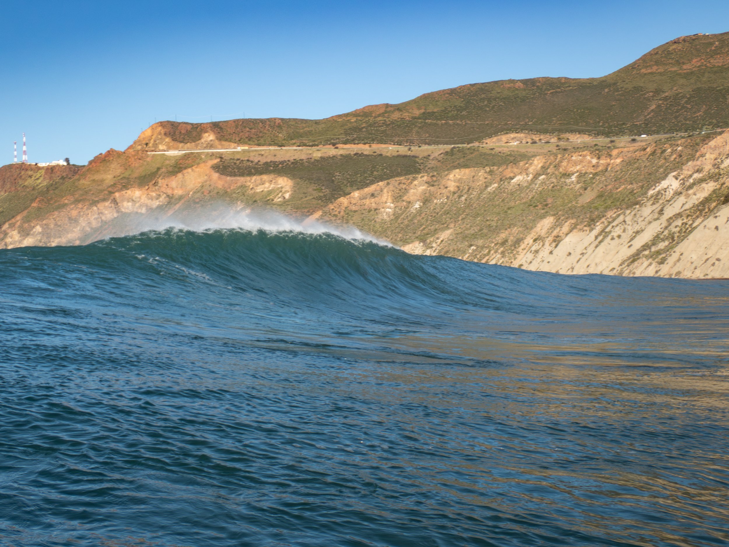 Large ocean wave with a hilly coastline and clear blue sky in the background.