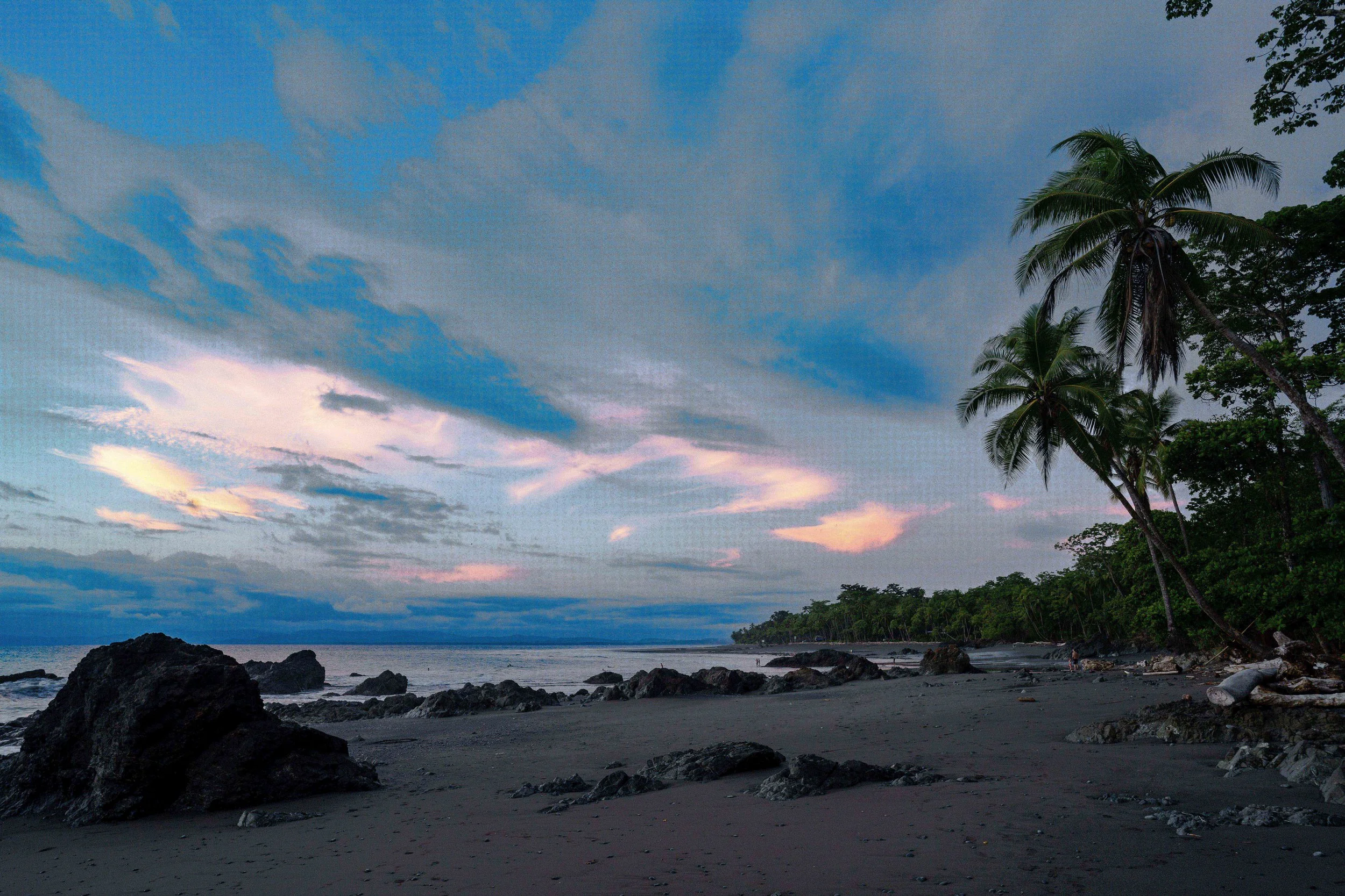 Beach at dusk with rocks on the shore, tropical trees, and a sky with clouds and pink and purple hues.
