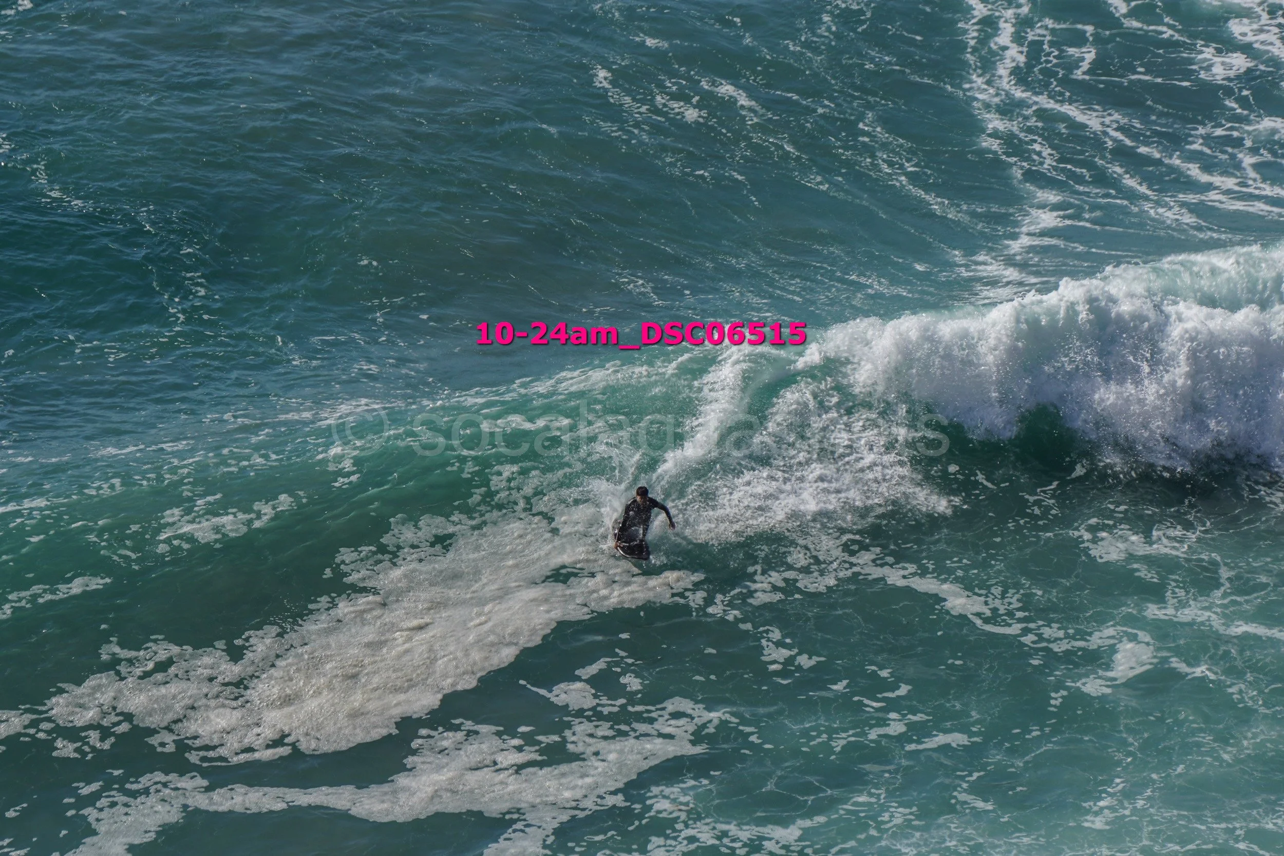 A person surfing on a large ocean wave with white foam, seen from above.