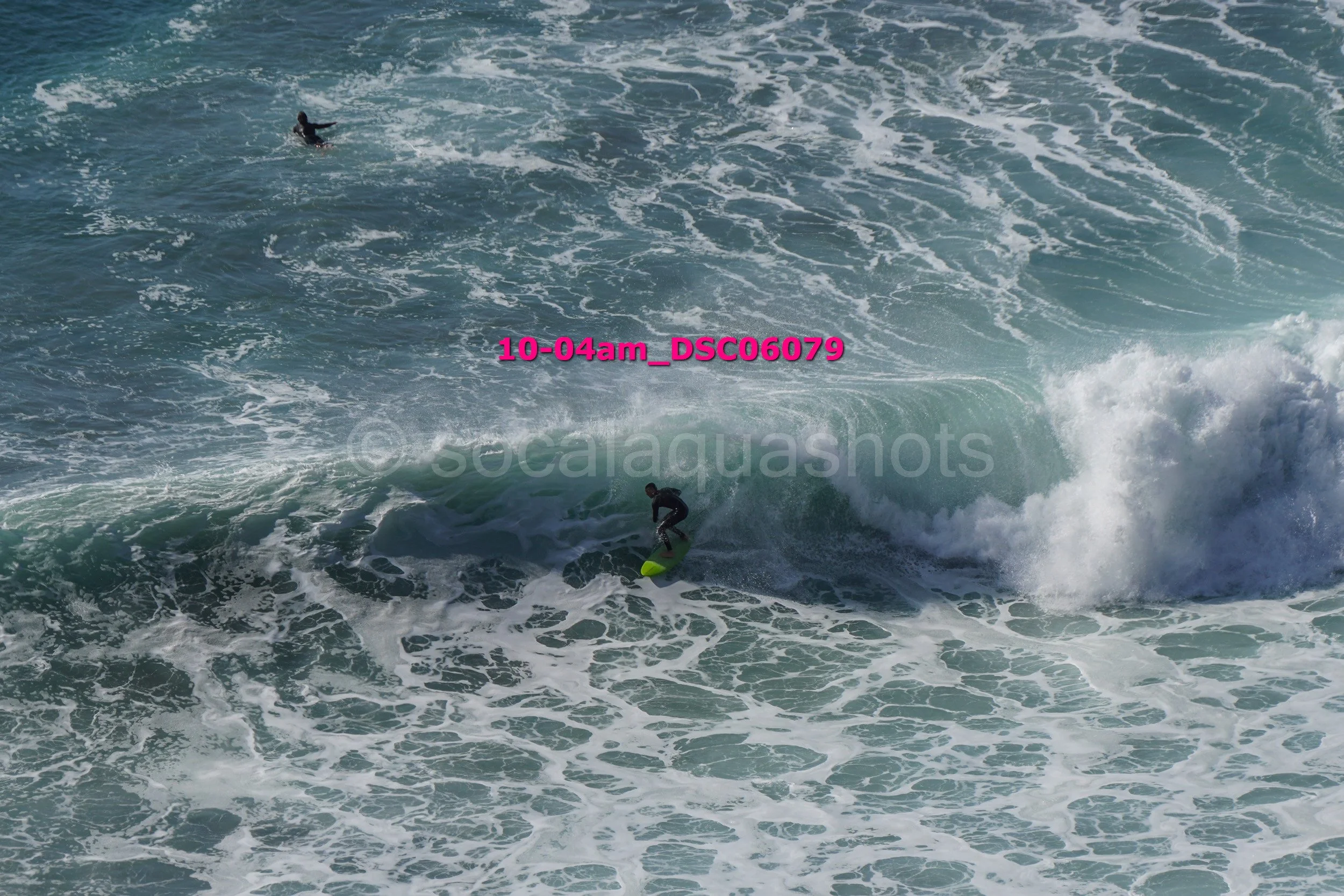A person surfing on a wave in the ocean with another swimmer in the water, seen from above during daytime.